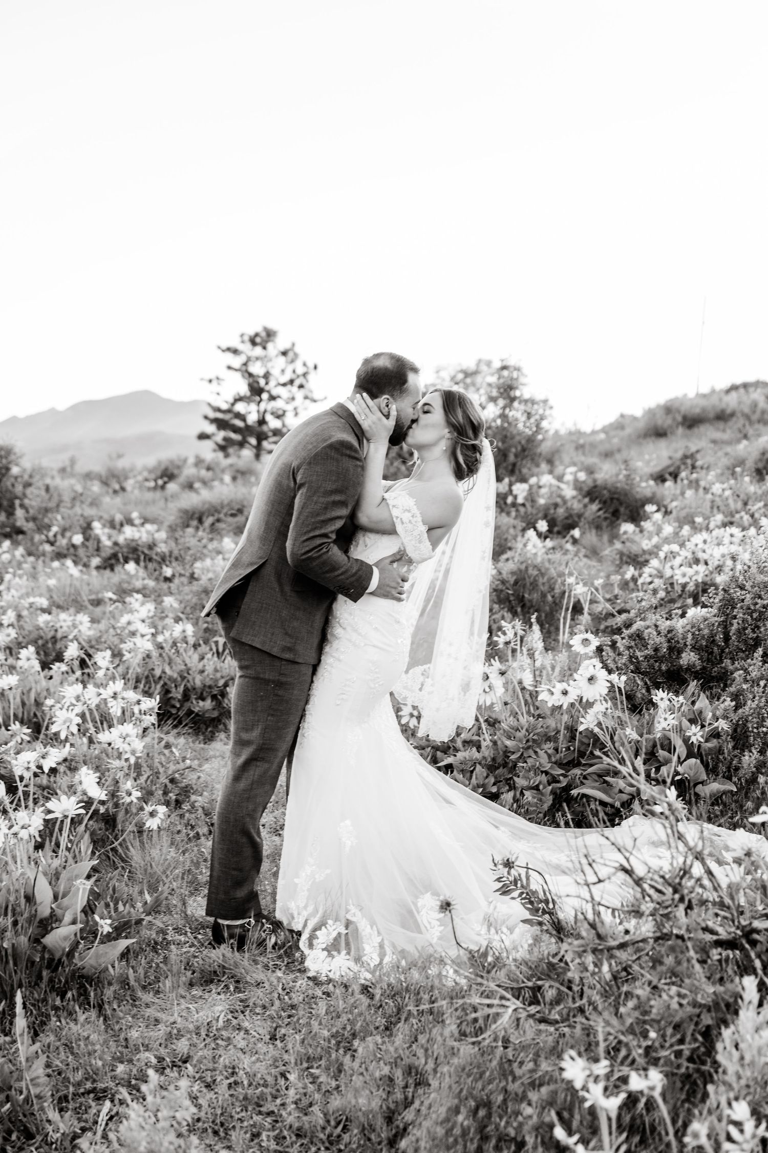 A romantic wedding couple embraces in a field of wildflowers with mountains in the background.
