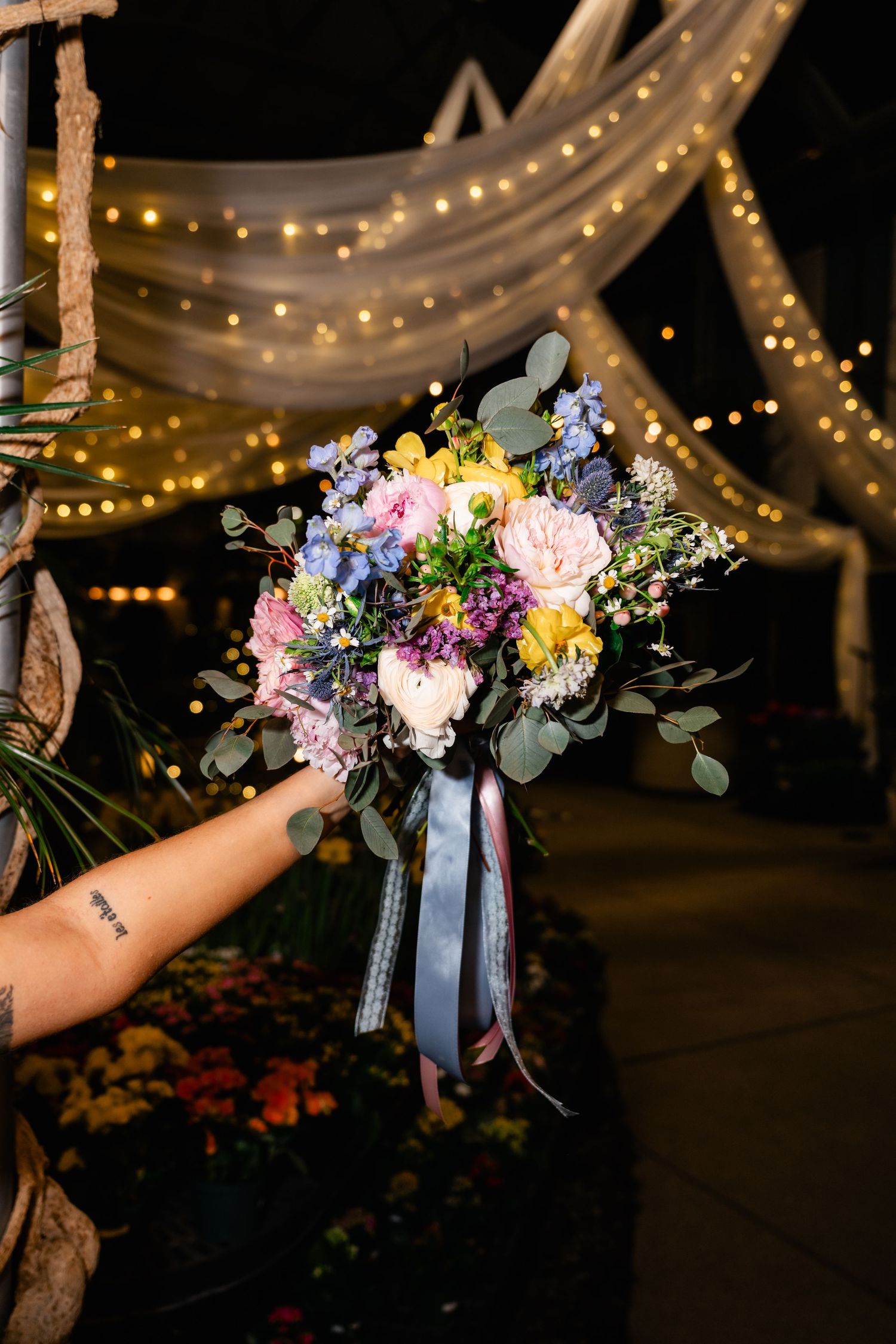 A colorful wedding bouquet with pink, purple and yellow flowers held up against a background of twinkling string lights.