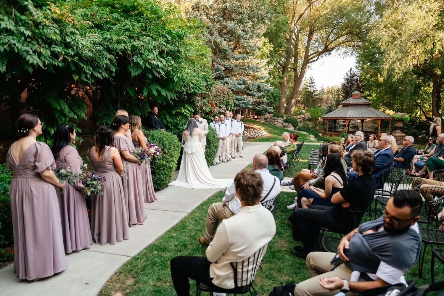 An outdoor garden wedding ceremony with bridesmaids in purple dresses and guests seated on white chairs.