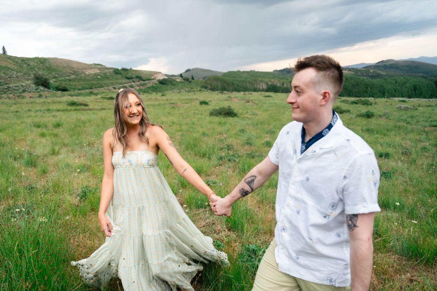A couple holds hands while walking through a green mountain meadow, with the bride wearing a flowing white dress. Guardsman Pass engagement