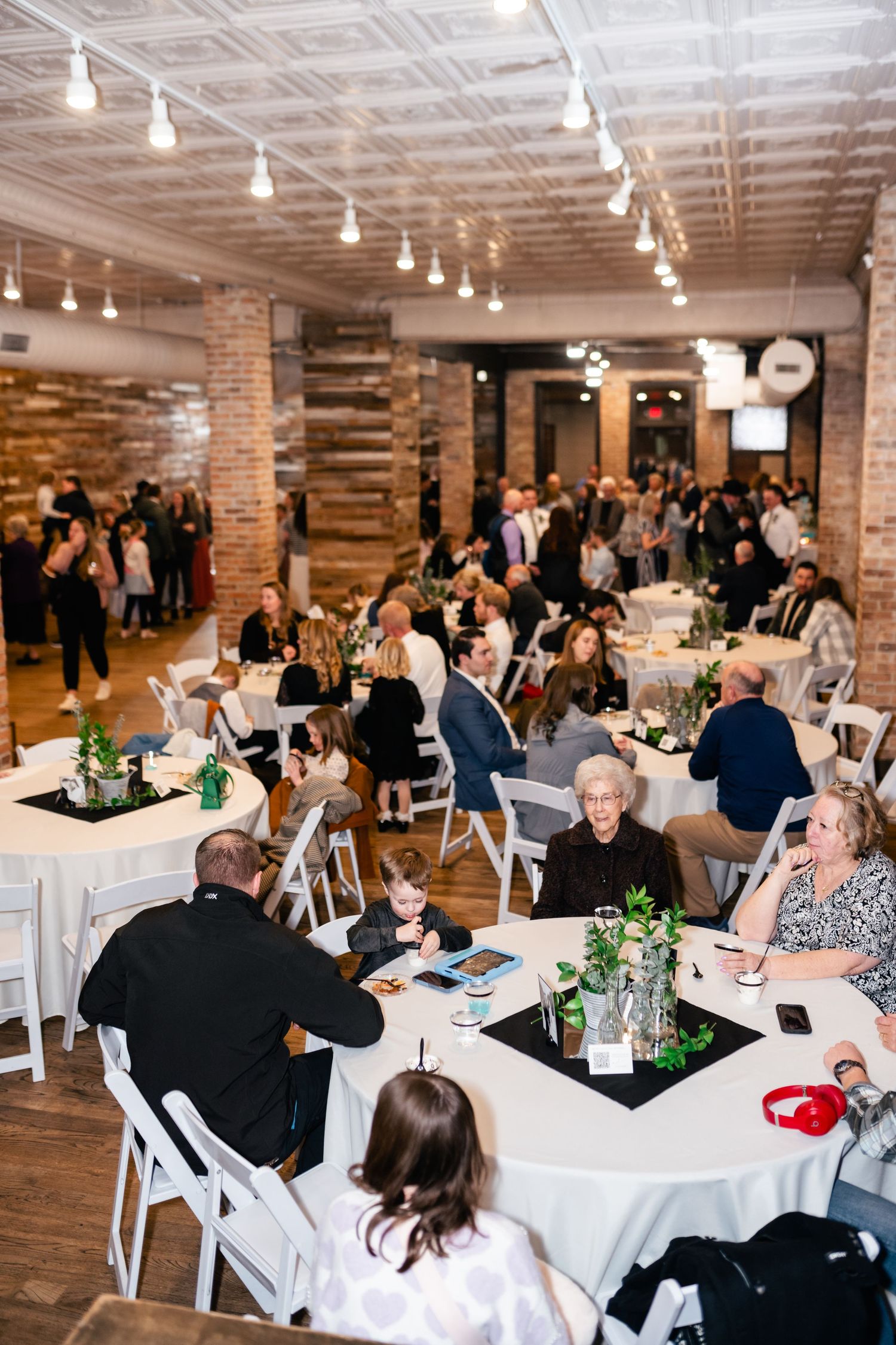 Wedding reception dining room with exposed brick walls, white tables, and industrial lighting overhead.