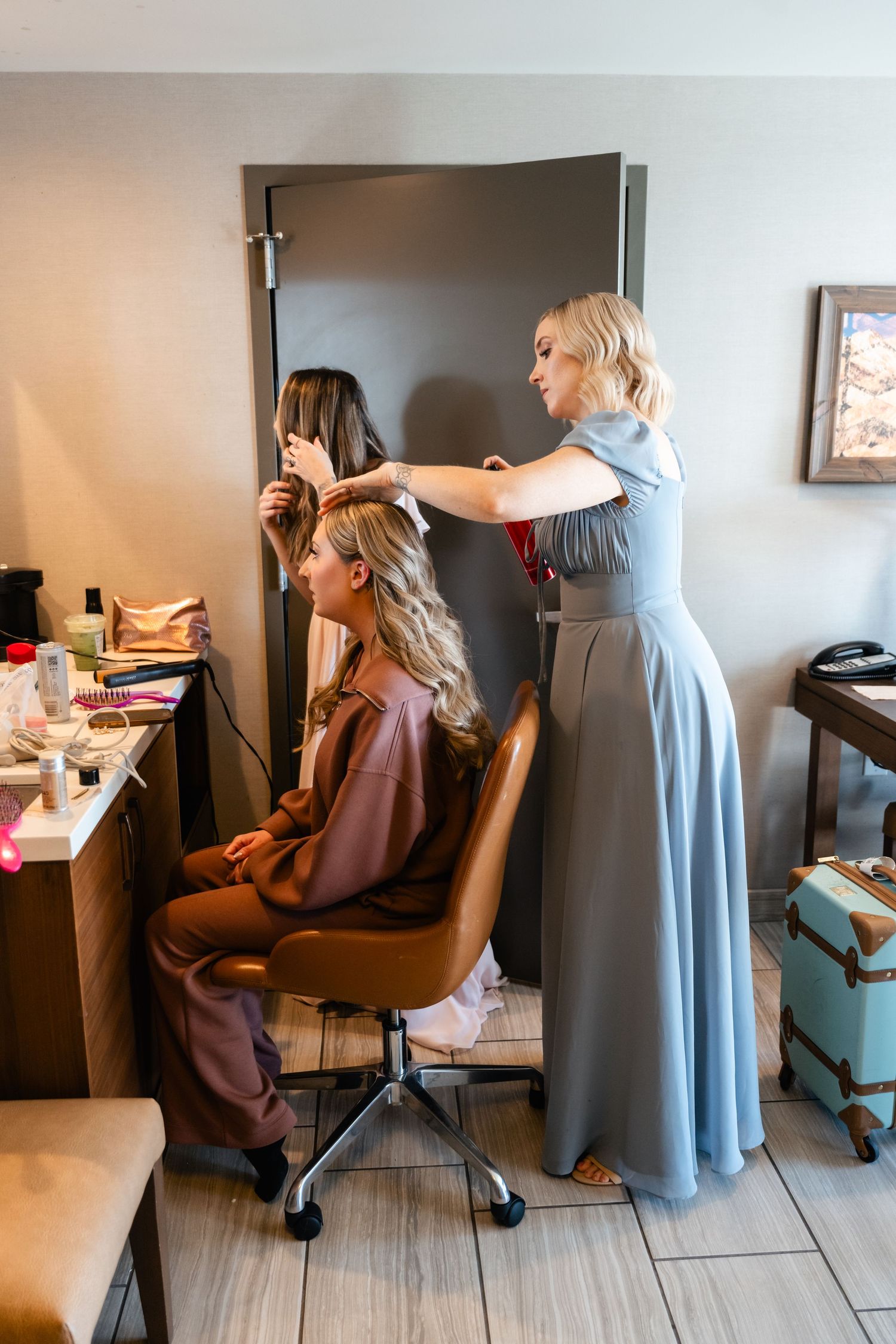 A stylist in a long blue dress works on styling a client's hair in a salon setting with beauty supplies on the counter.