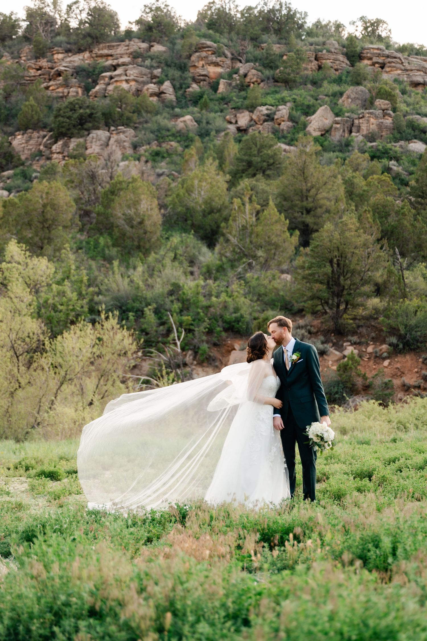 A romantic wedding photoshoot in a green field with dramatic mountain cliffs in the background.