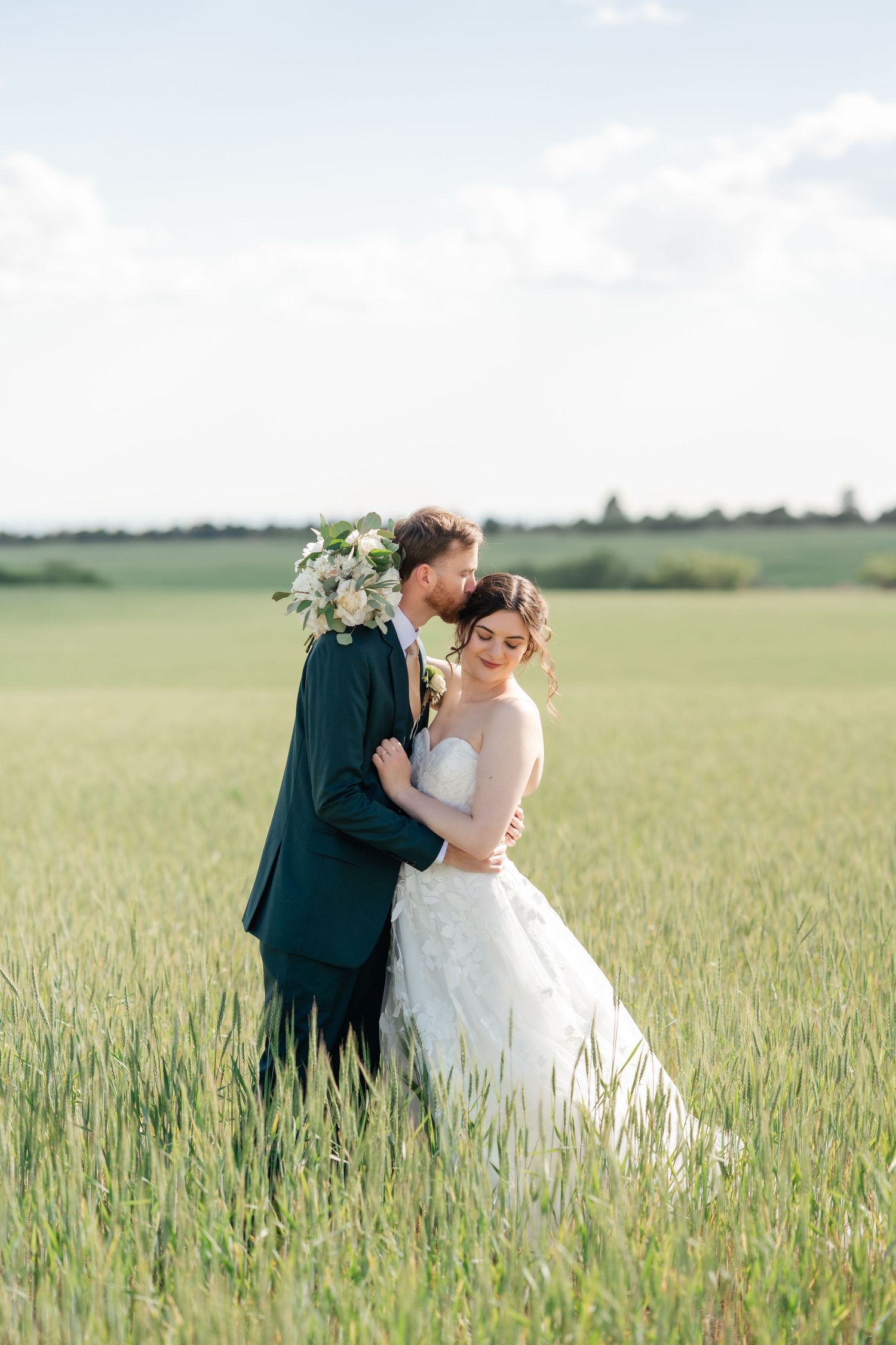 A romantic couple embraces in a sunlit wheat field wearing wedding attire during a countryside photoshoot.