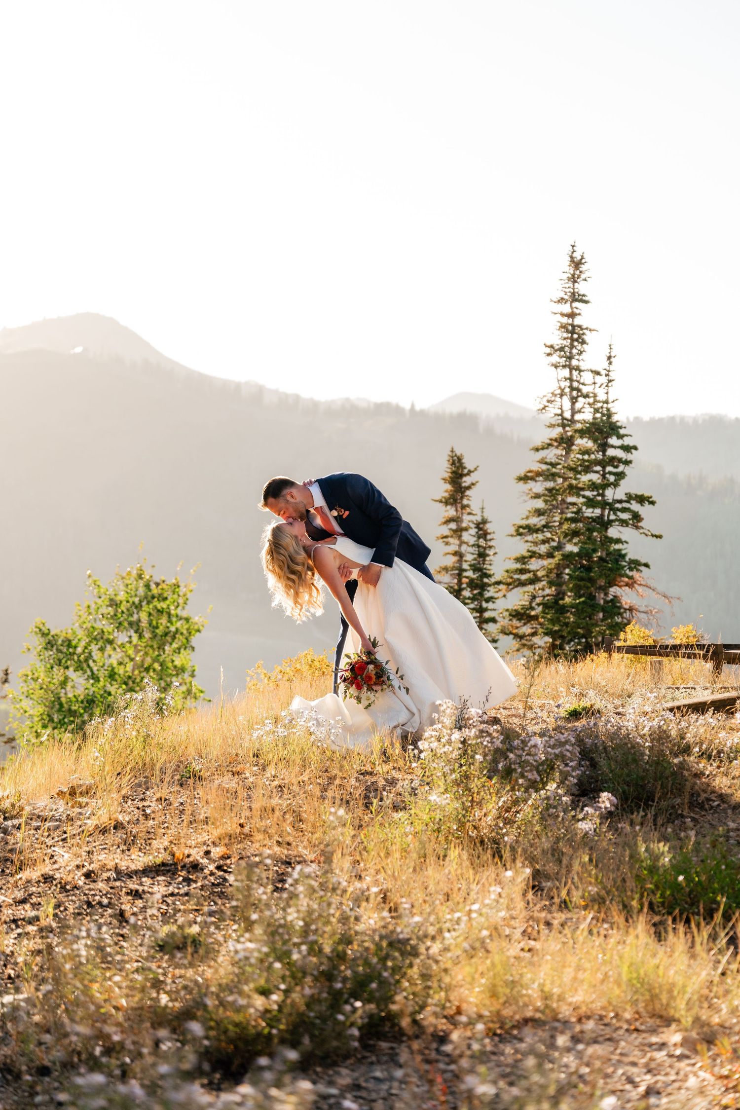 A romantic wedding photo shoot on a mountainside with golden sunlight and evergreen trees in the background.