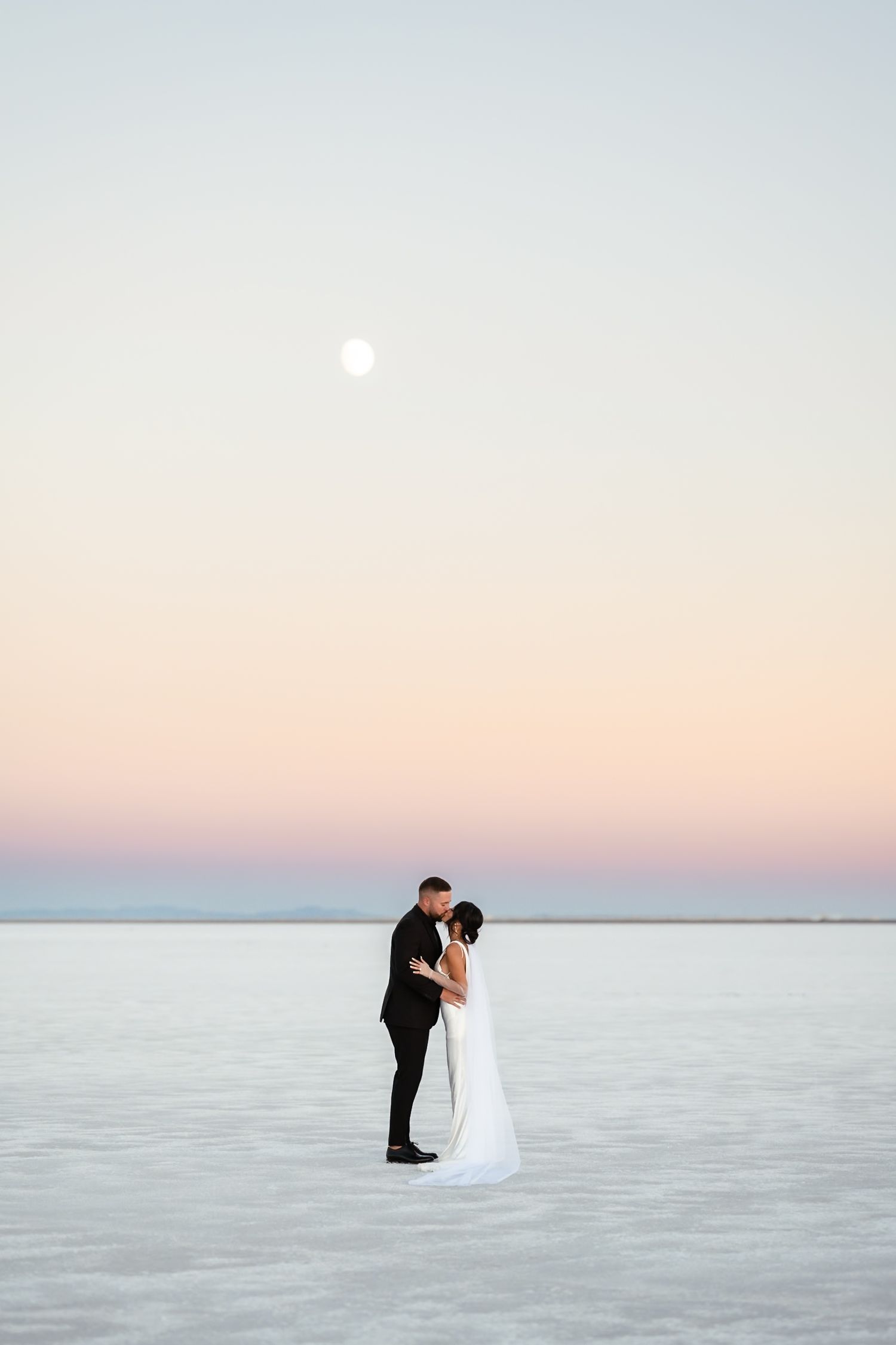 Wedding couple poses against pastel sky on salt flats with full moon visible.