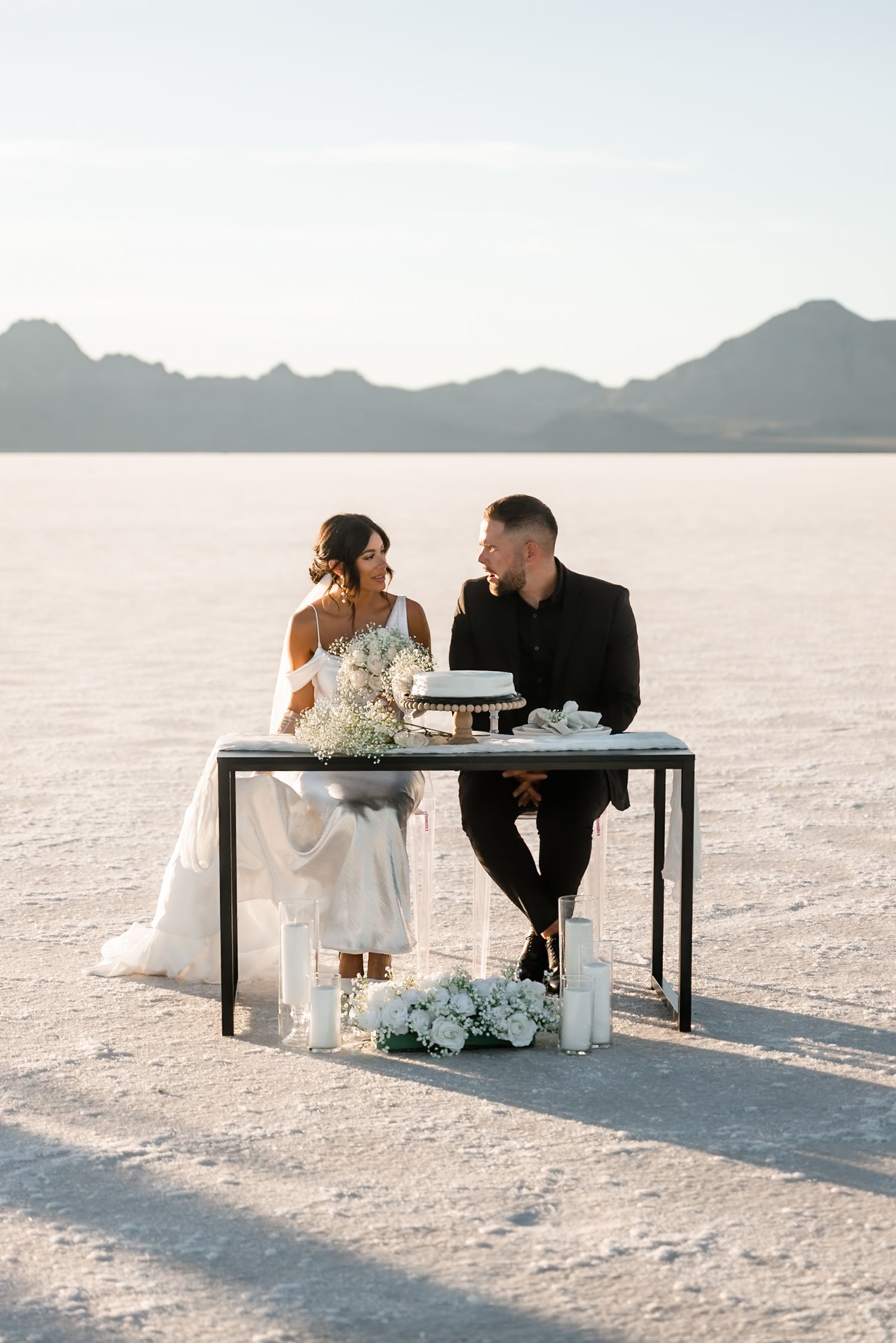 Newlyweds share drinks at a modern black table set up on the bright white salt flats.