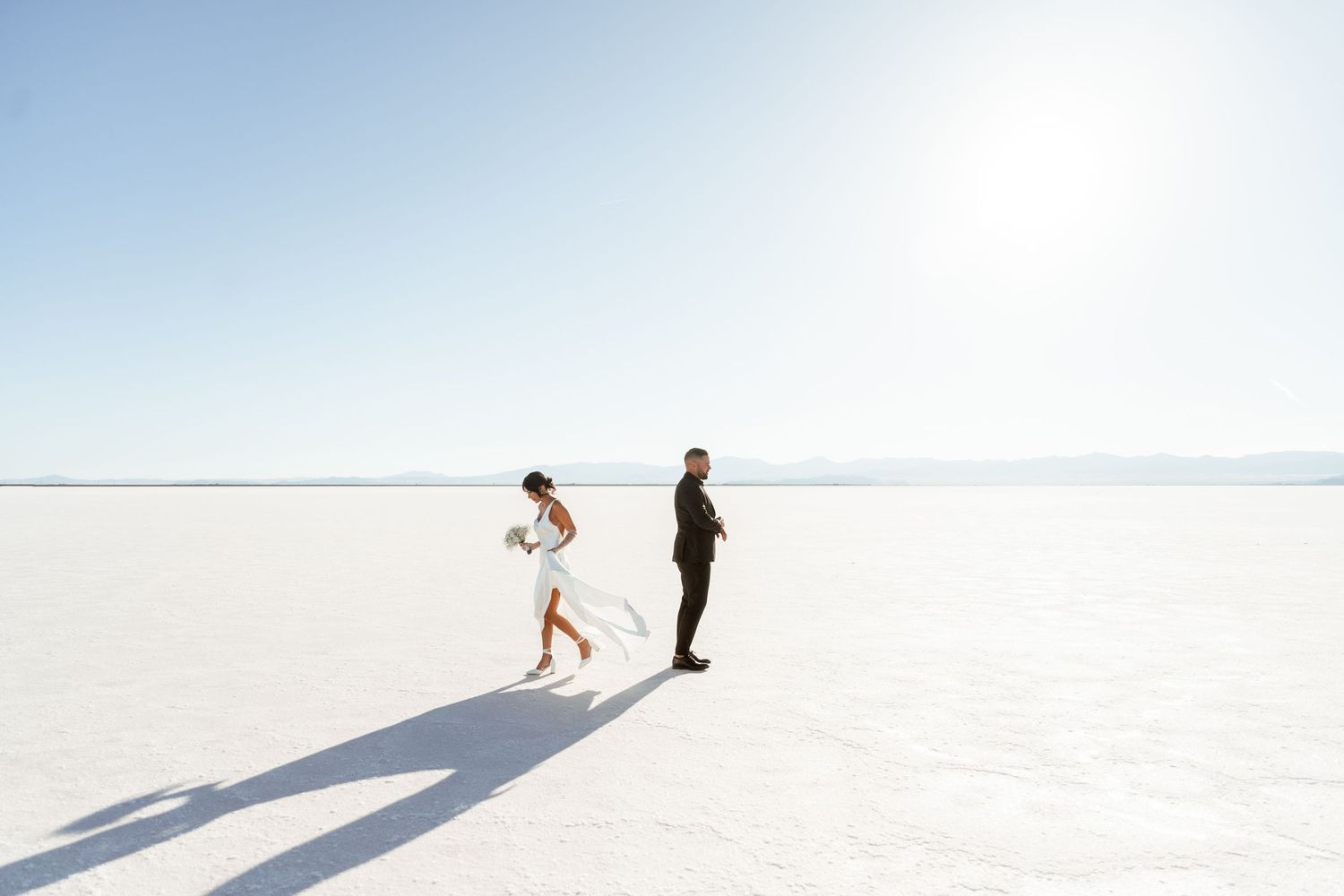 Two figures walking together on a vast white salt flat with long shadows cast behind them in bright sunlight.