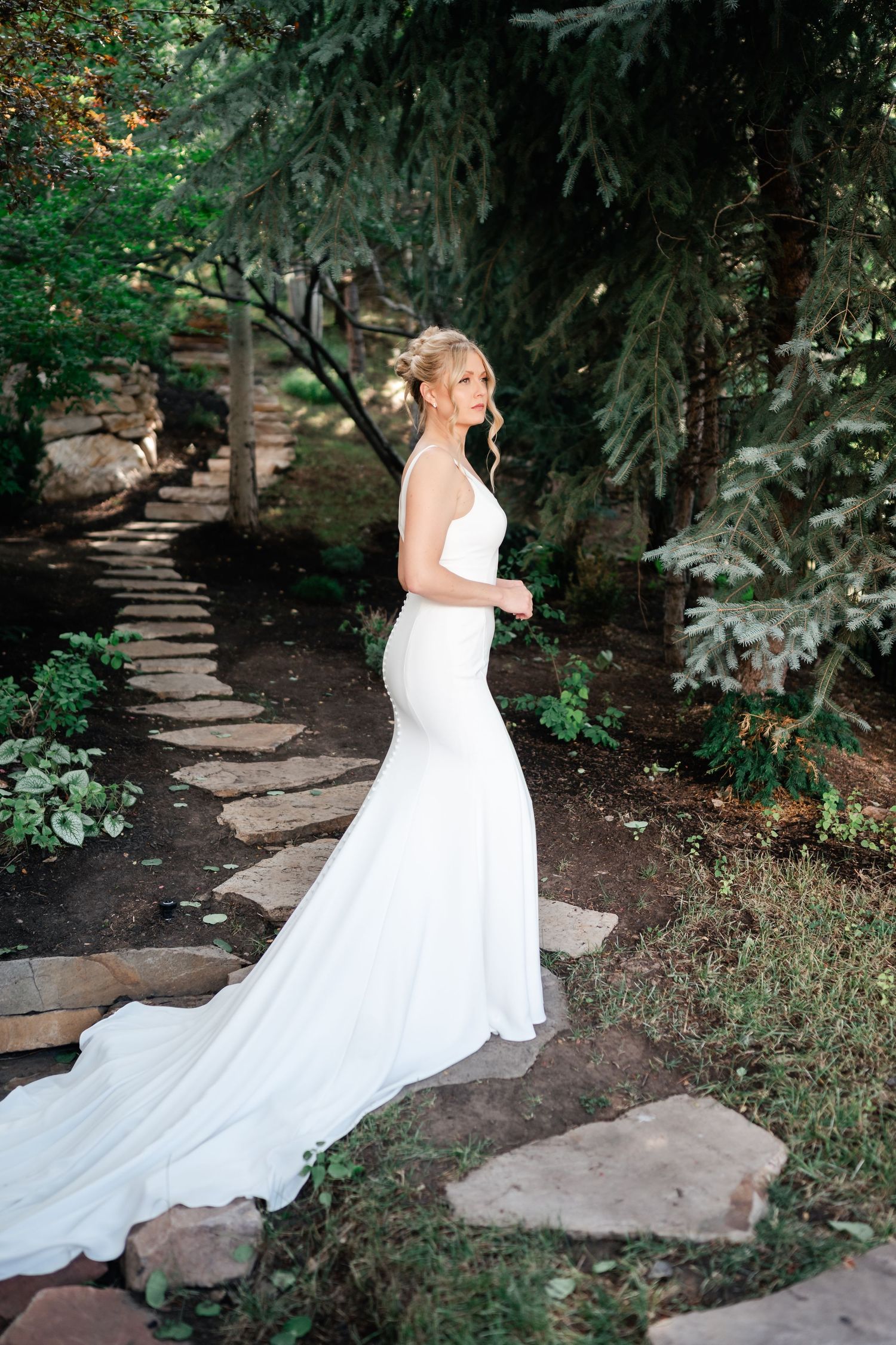 Bride in flowing white gown poses on stone pathway surrounded by lush garden.