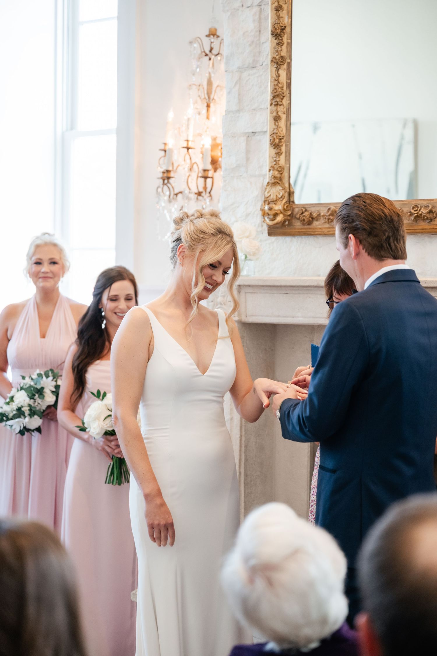 An intimate wedding ceremony takes place in an elegant room with a crystal chandelier and ornate gold mirror.