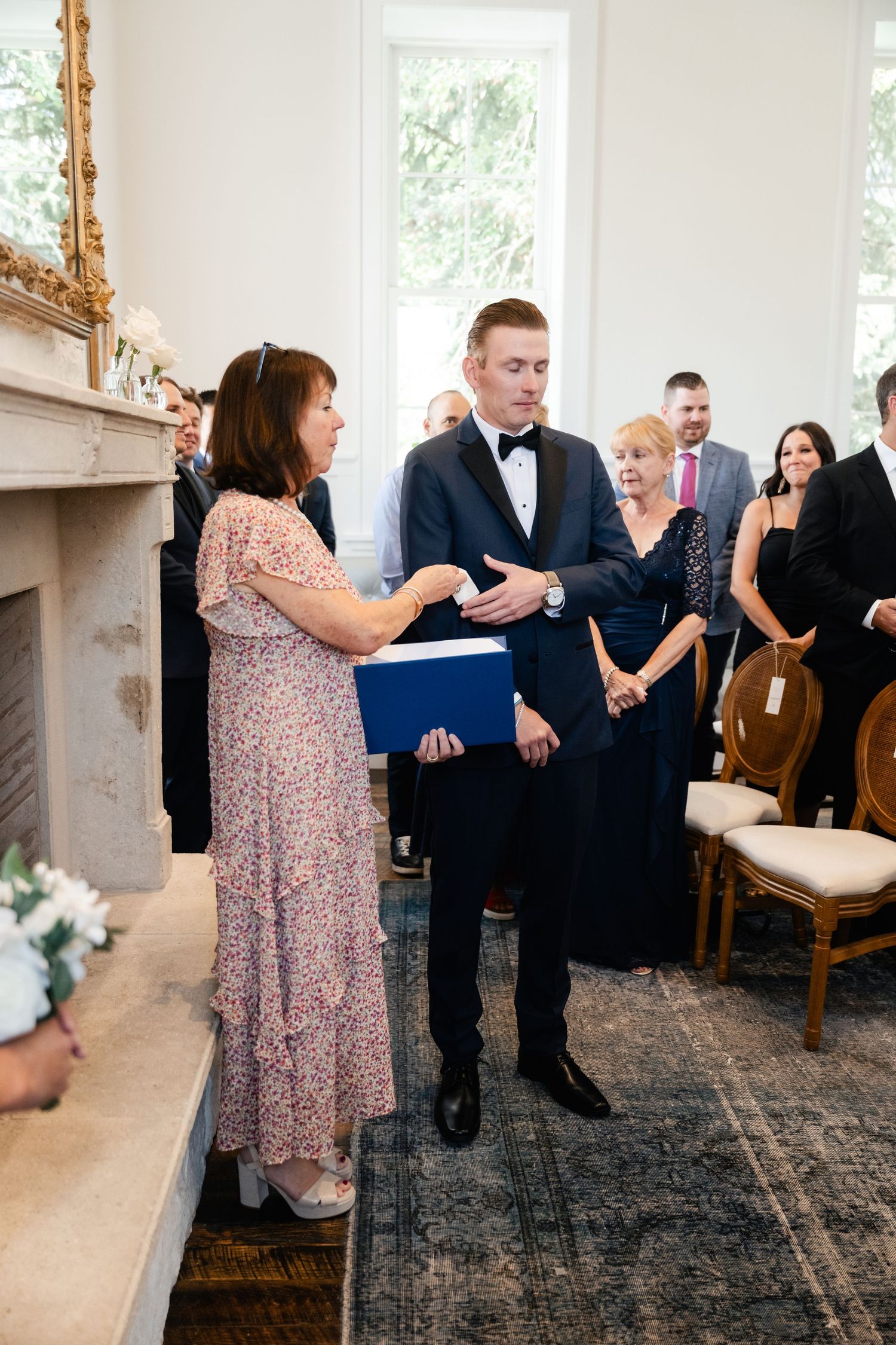 Wedding guests share an emotional moment during a reception in an elegant white room with hardwood floors.
