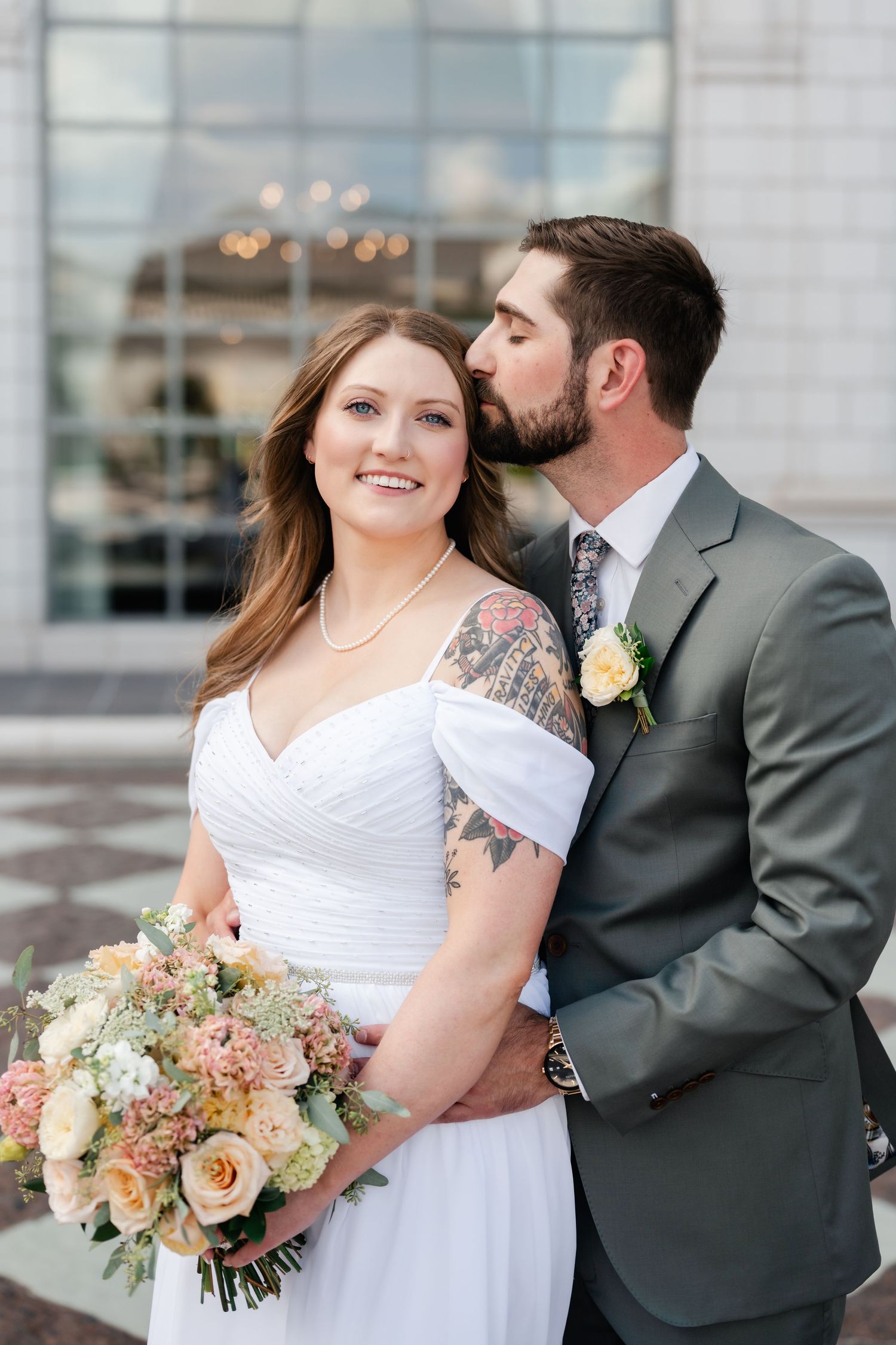 A bride holding a peach and white bouquet poses with her groom in front of a modern building.