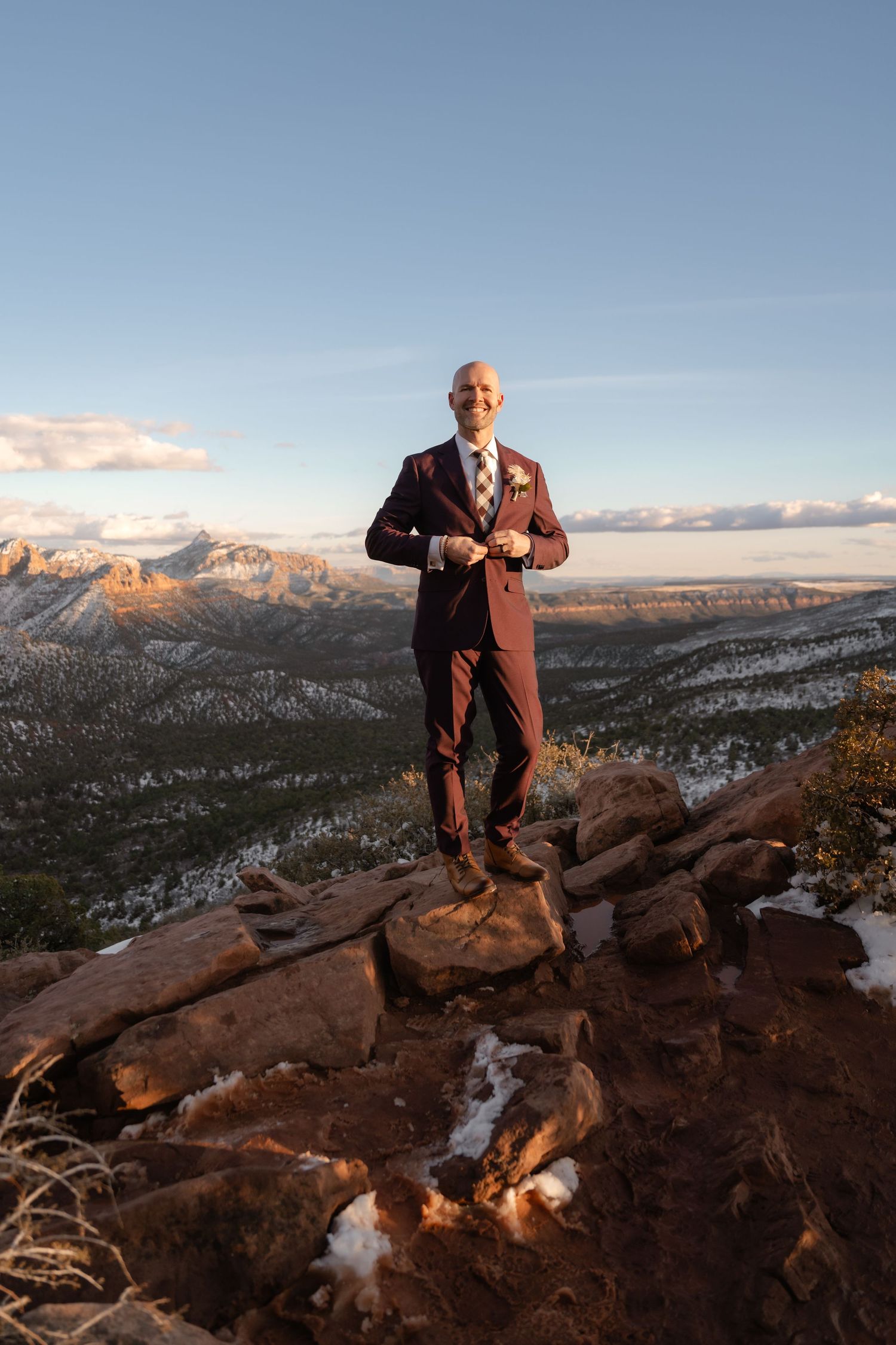 A person in formal attire poses on red rocks overlooking a snow-dusted mountain valley.