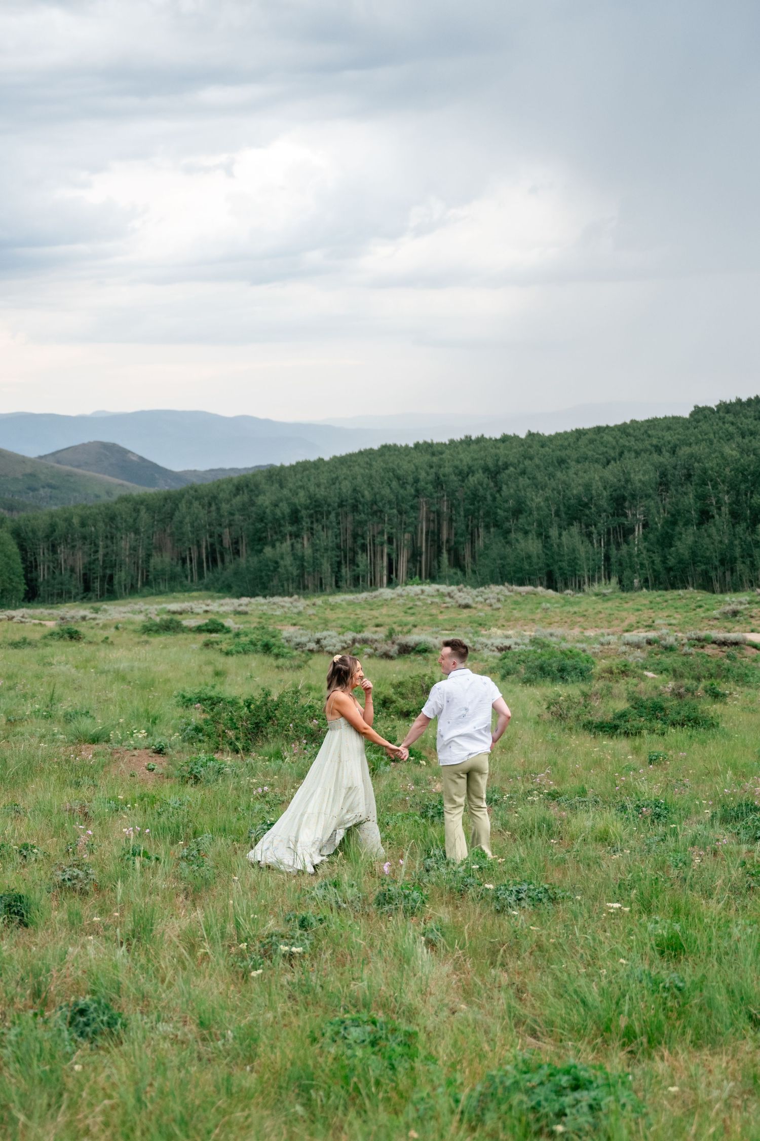 A couple stands together in a vast mountain meadow with forest and peaks in the background.