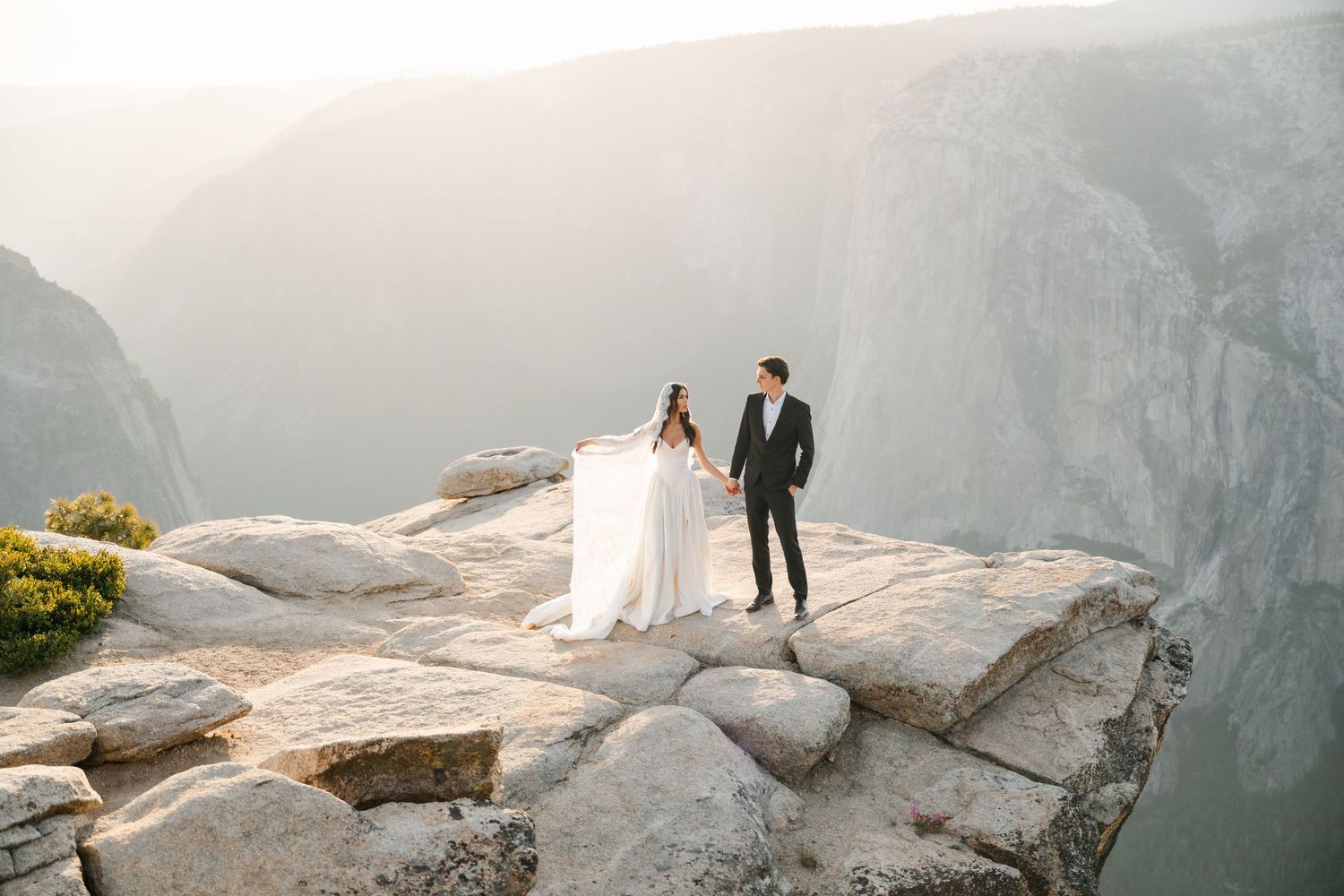 Bride and groom holding hands on rocky Yosemite cliff edge with dramatic canyon and hazy mountains.