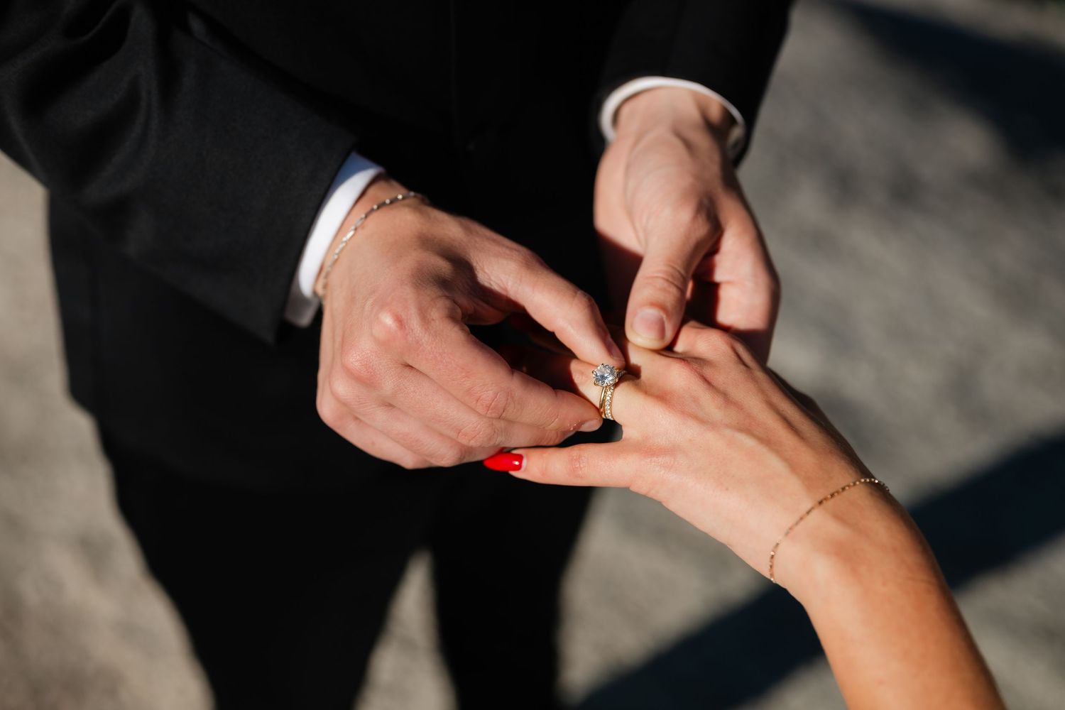 Groom placing a diamond engagement ring on bride's finger with red nails and gold bracelet during wedding ceremony.