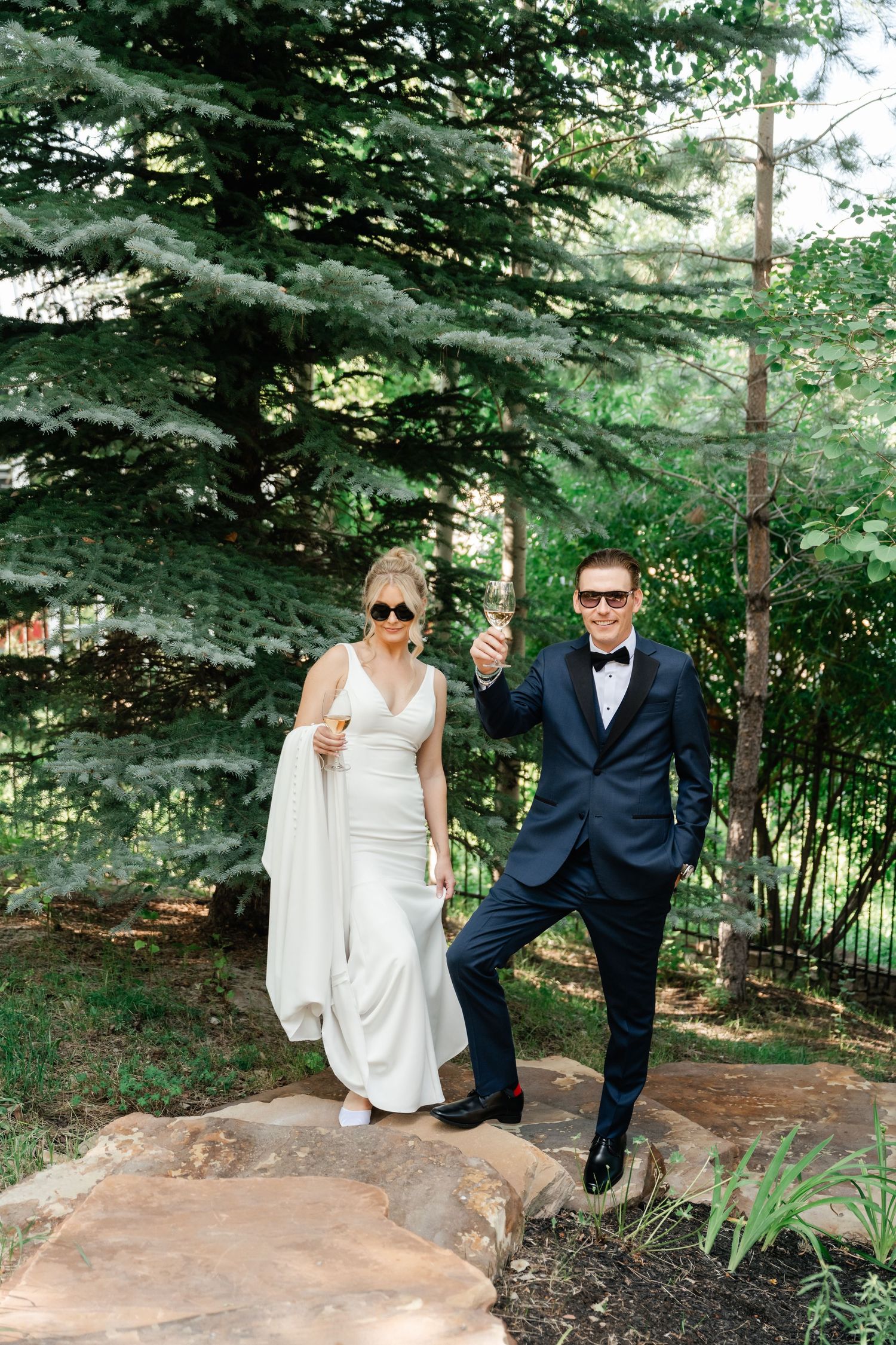 Bride in white gown and groom in navy tuxedo toasting with champagne outdoors among tall pine trees at their intimate Park City wedding.