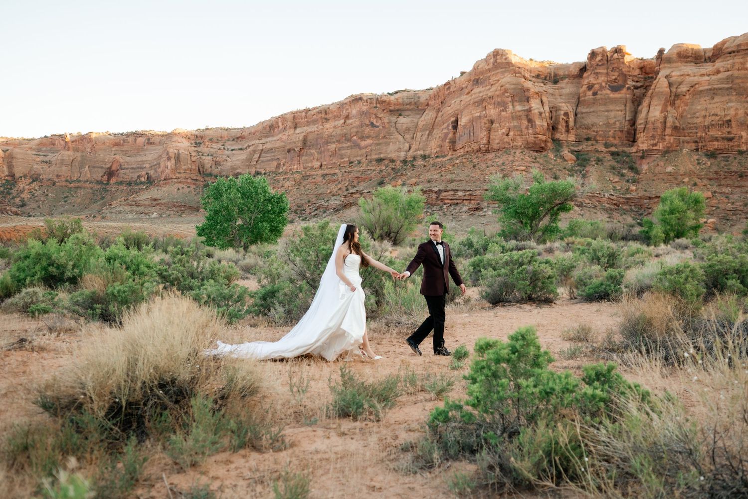 Bride and groom walk hand-in-hand through desert landscape with red rock formations in background.