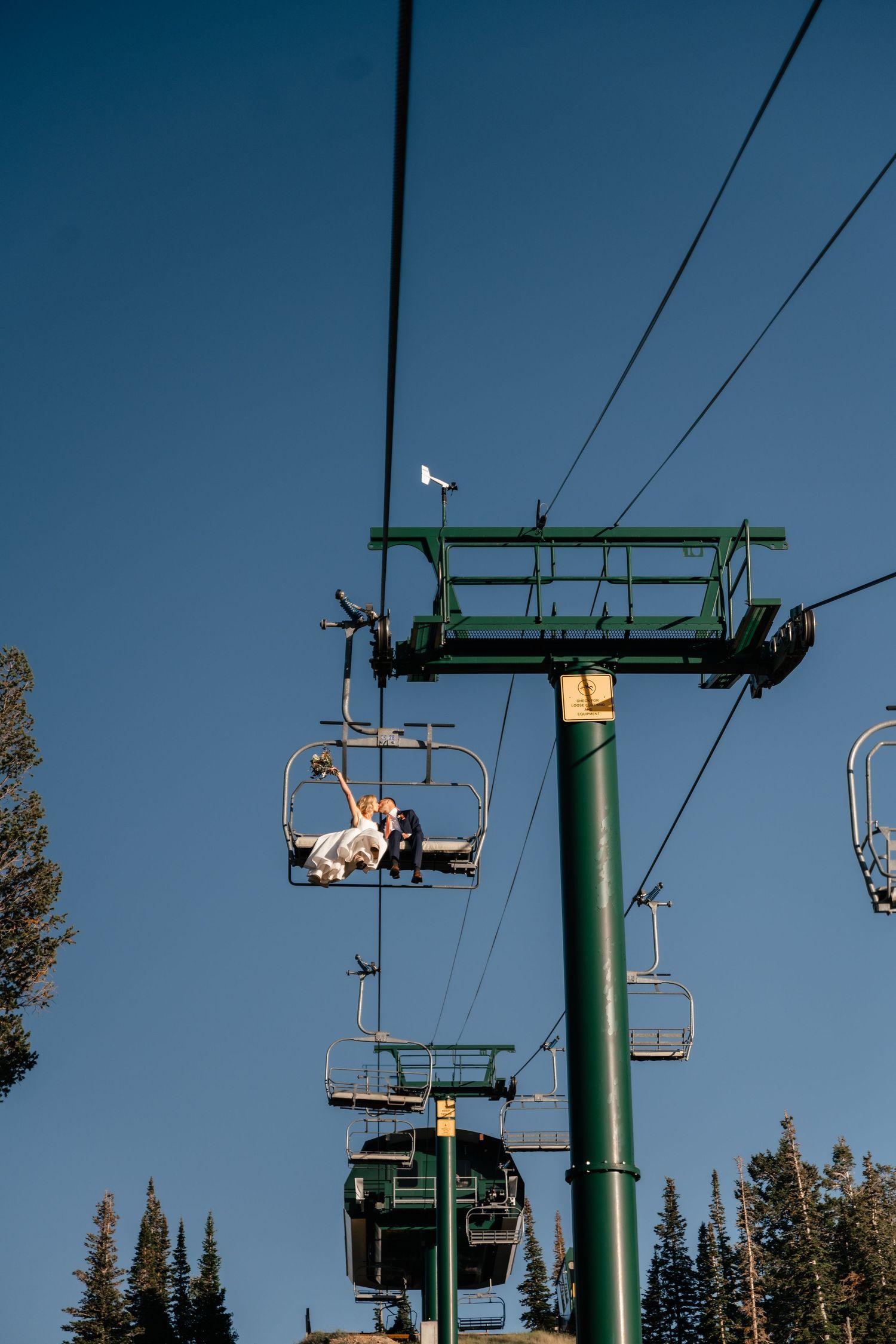 A ski lift chair rises against a bright blue sky with evergreen trees and mountain scenery in the background.