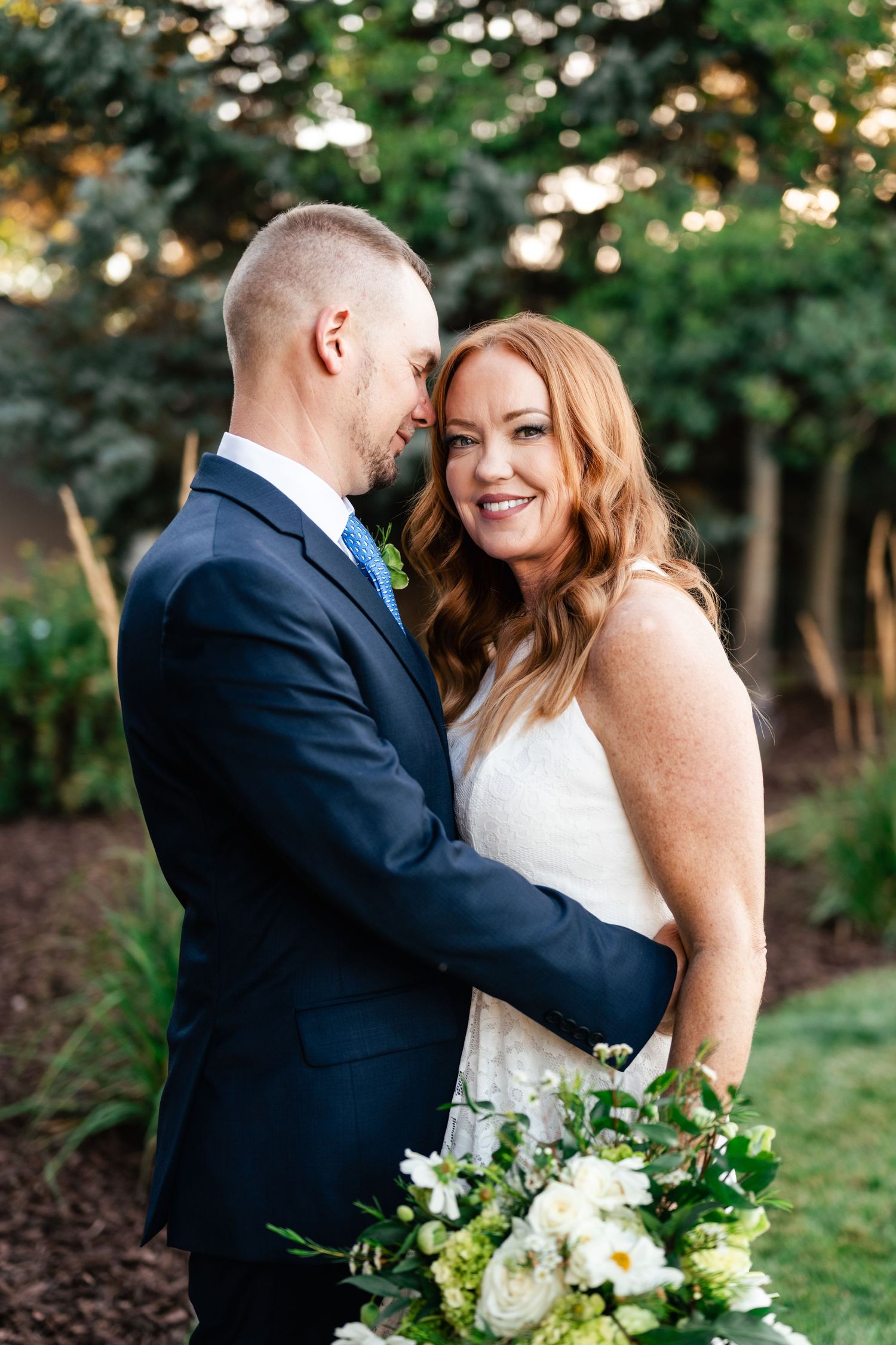Newlywed couple shares intimate moment in garden setting with white floral bouquet.