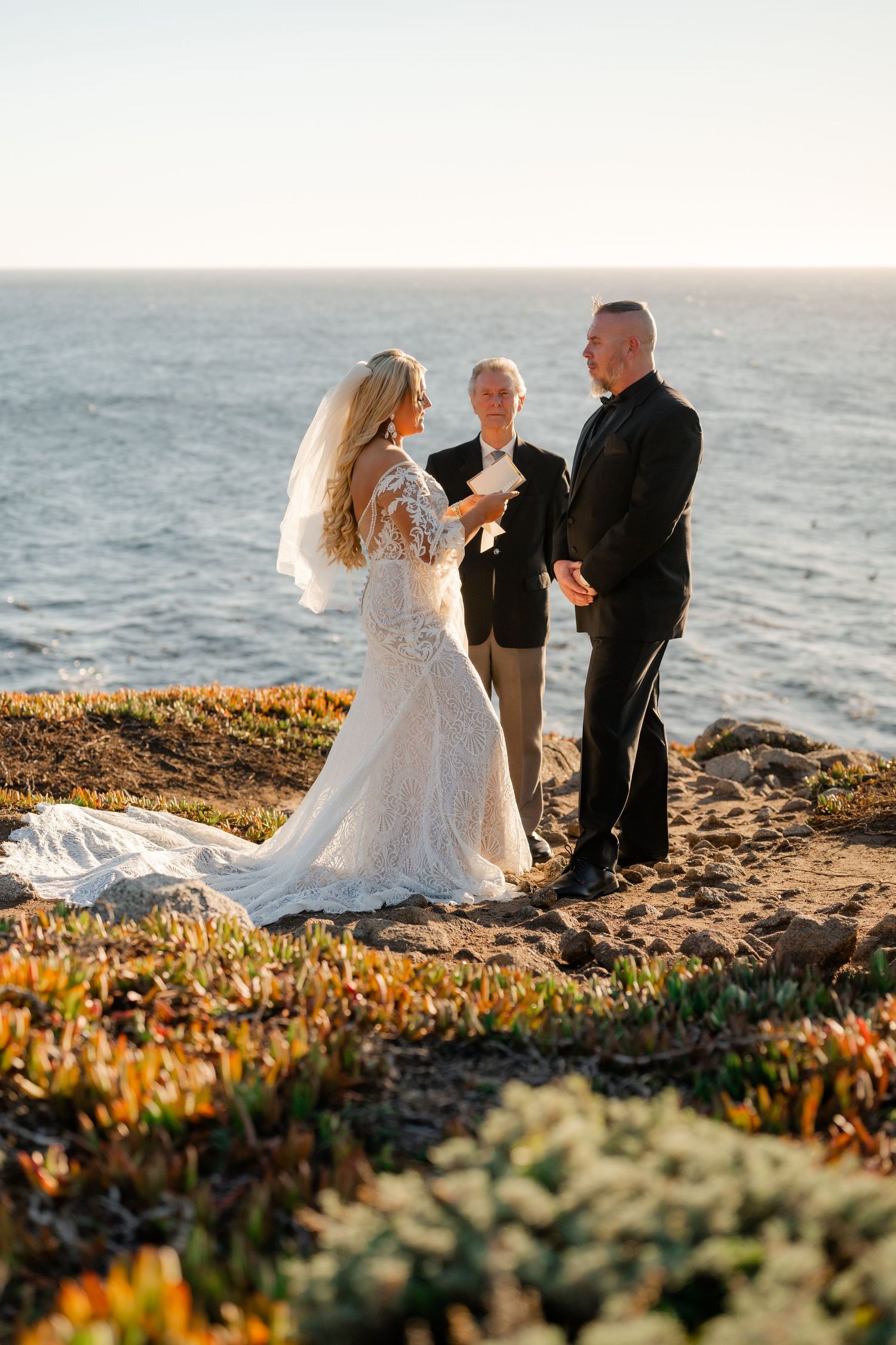 Seaside wedding ceremony unfolds against dramatic Pacific coast landscape with golden vegetation.