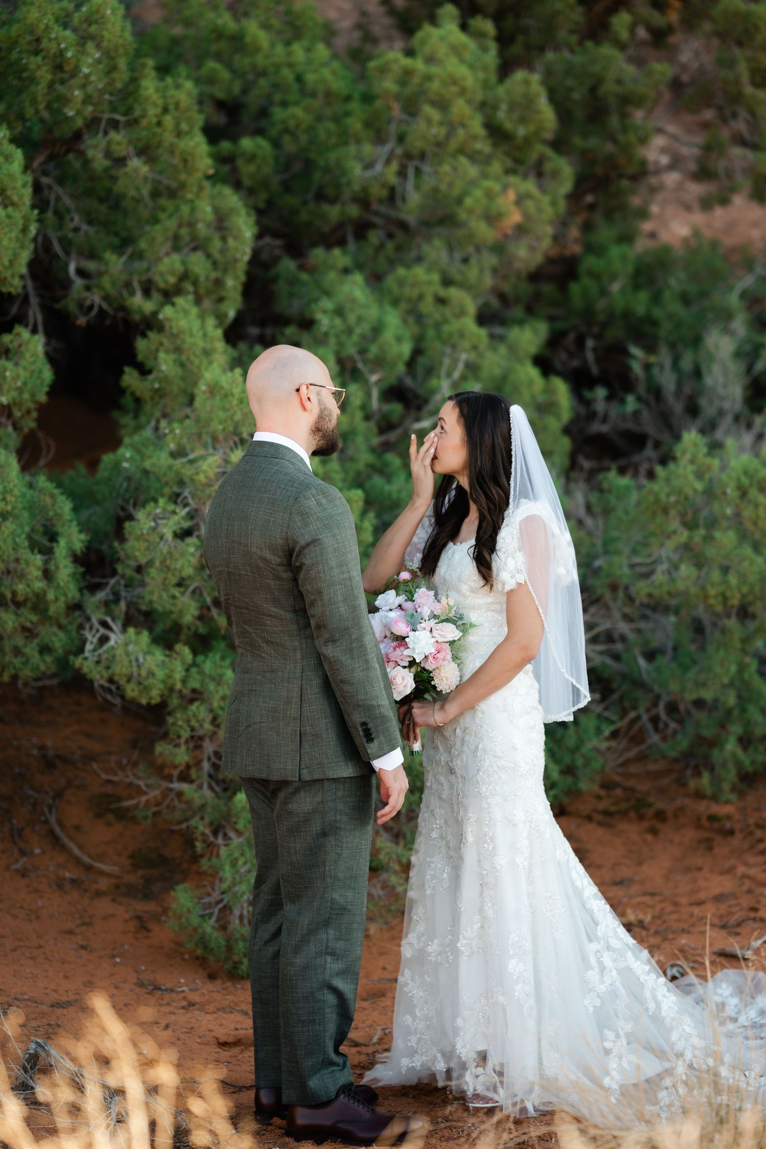 A romantic wedding moment captured among green desert shrubs with red rocks in the background.