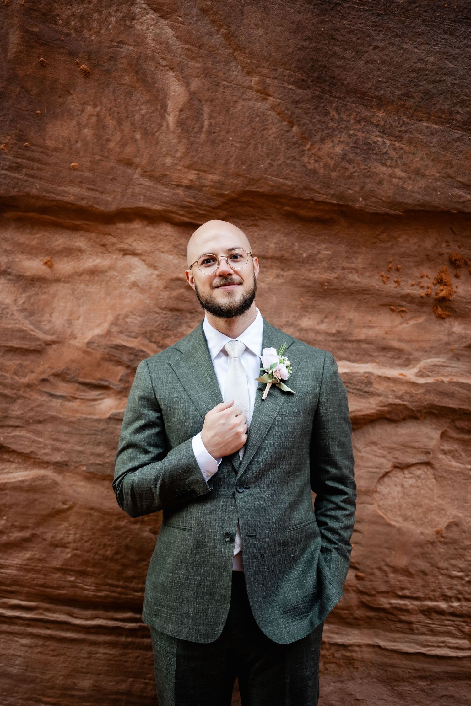 A groom in a green suit adjusts his tie while standing against red rock formations.