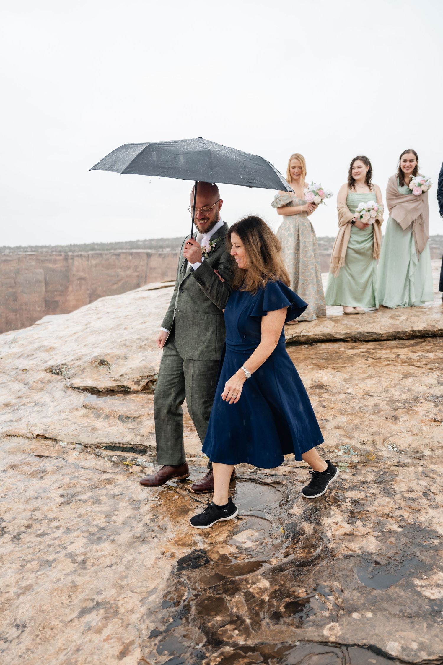 Wedding party members in formal attire gather on rocky desert terrain under umbrellas.