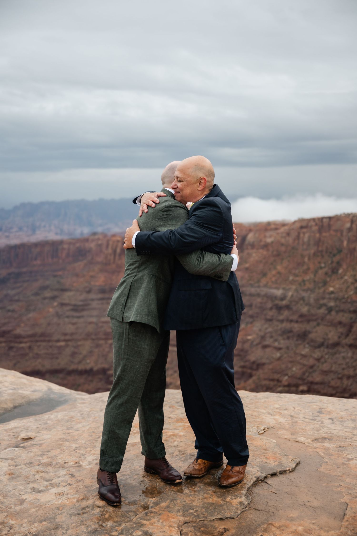 Two people share an emotional embrace on the edge of a dramatic canyon overlook with red rock cliffs in the background.