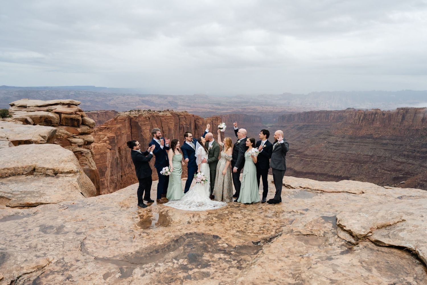 Wedding party stands along canyon rim with sweeping desert vista and dramatic cliffs in background.