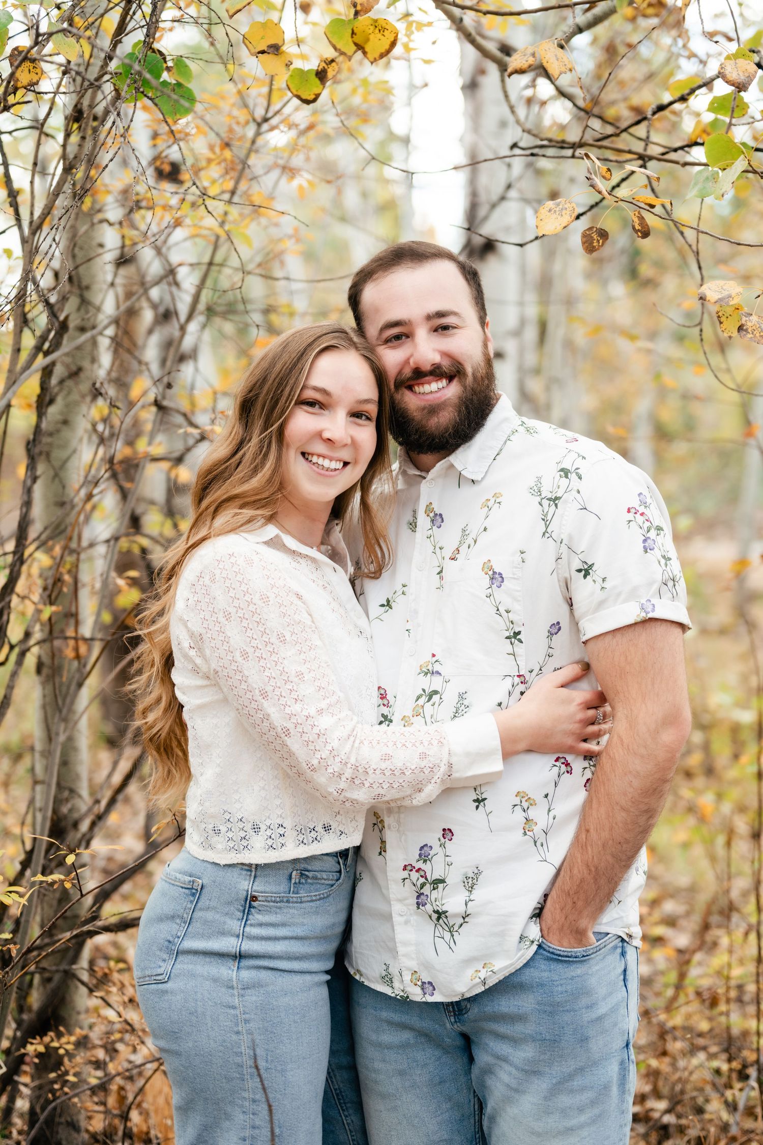 A couple poses playfully together in an autumn forest setting with yellow leaves and trees in the background. Engagement Photographer.