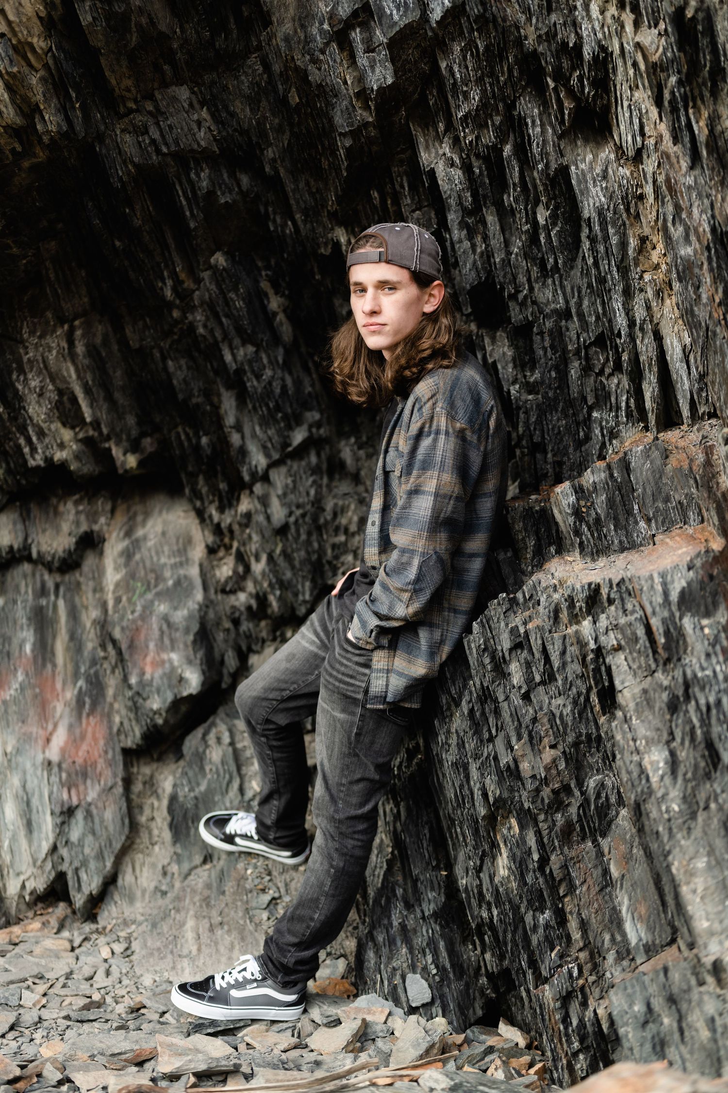 Person in plaid shirt and backwards cap poses against dark rocky cliff wall wearing black jeans and sneakers.