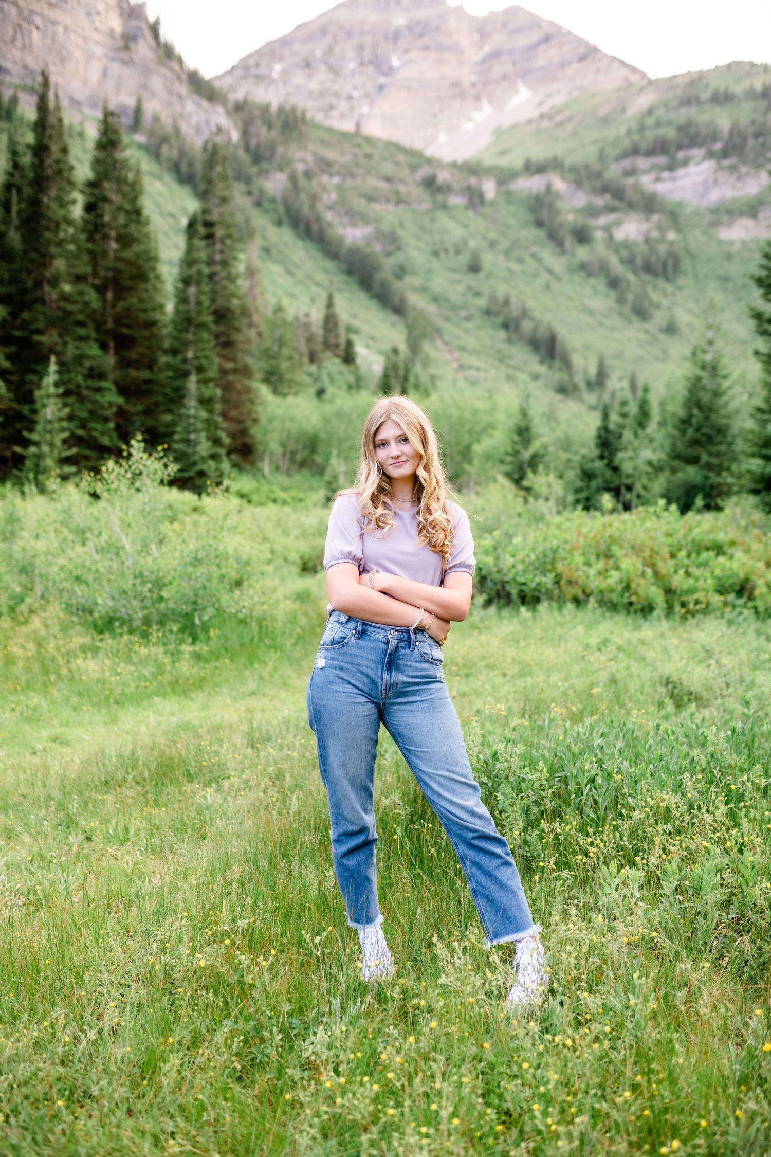 A person in jeans and white sneakers poses in a scenic mountain meadow filled with wildflowers and evergreen trees.