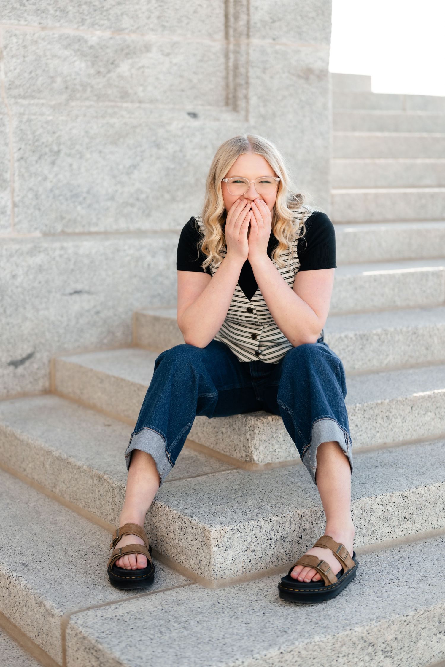 A person in a striped shirt and jeans sits on concrete steps wearing brown sandals while covering their mouth in surprise.