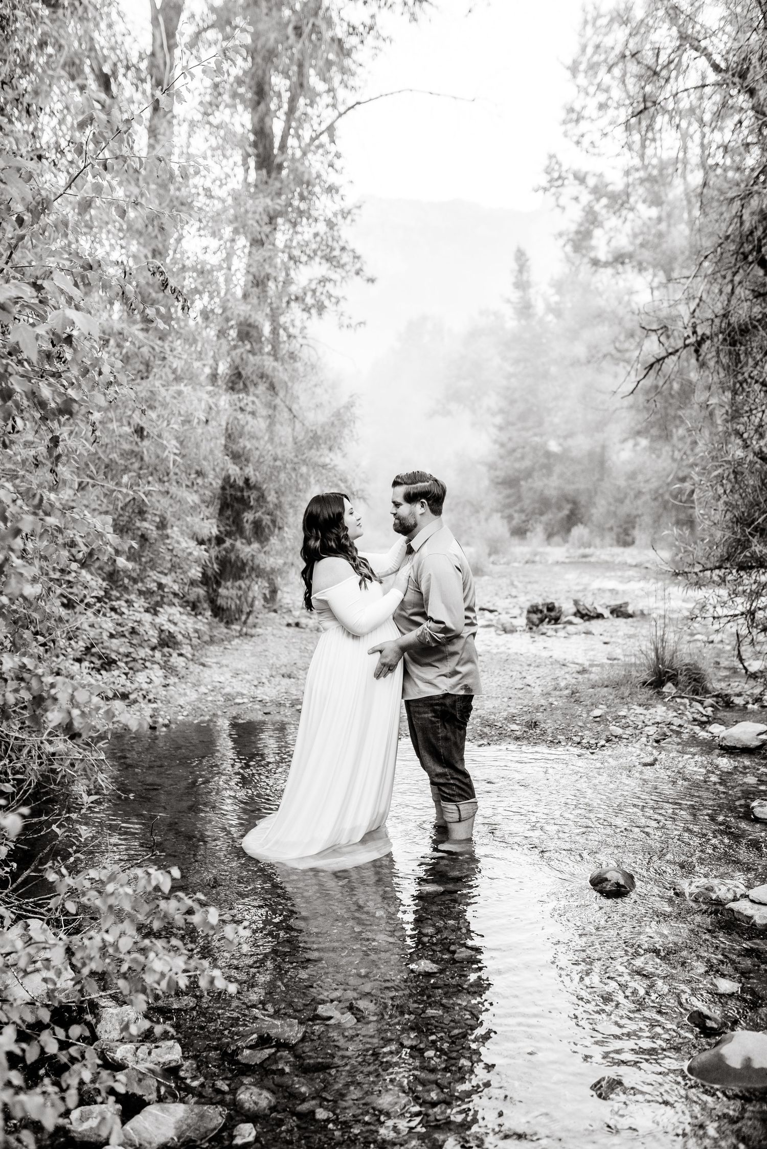 A couple stands together in a serene creek surrounded by trees in a series of artistic black and white photographs.