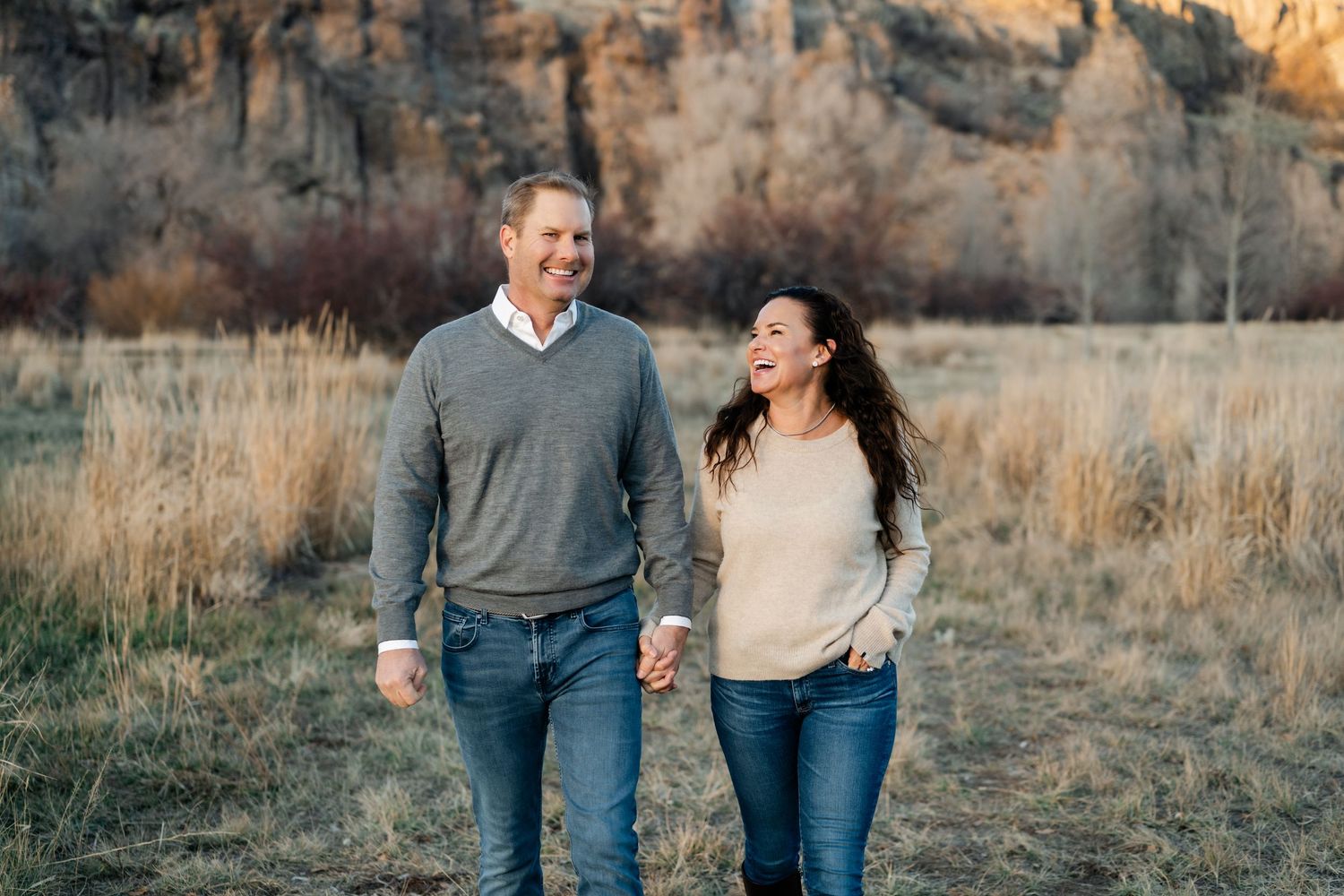 A couple walking hand in hand through tall autumn grass with rocky cliffs in the background at sunset.