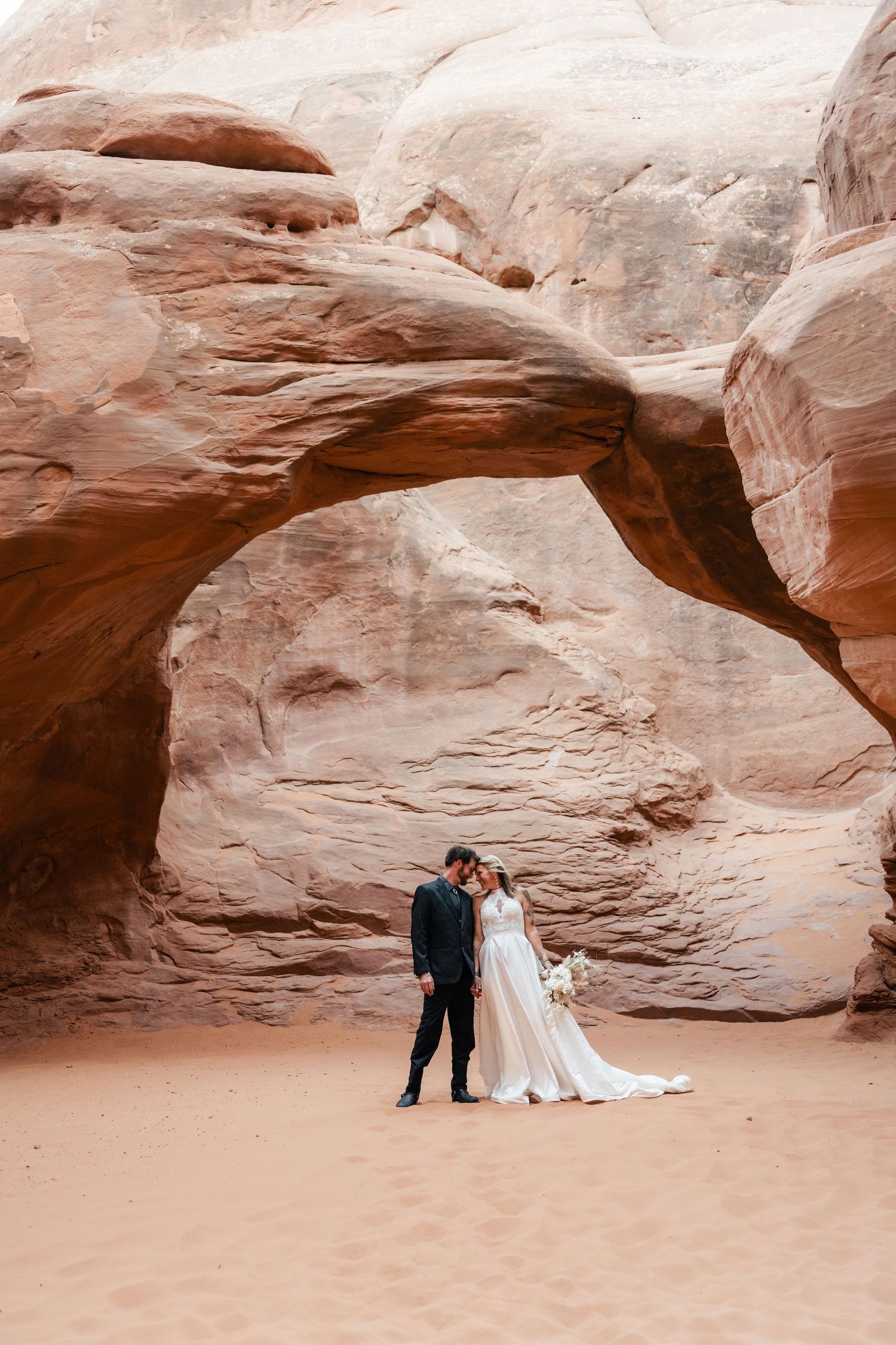 A couple shares an intimate moment under dramatic red rock arches in a desert landscape.