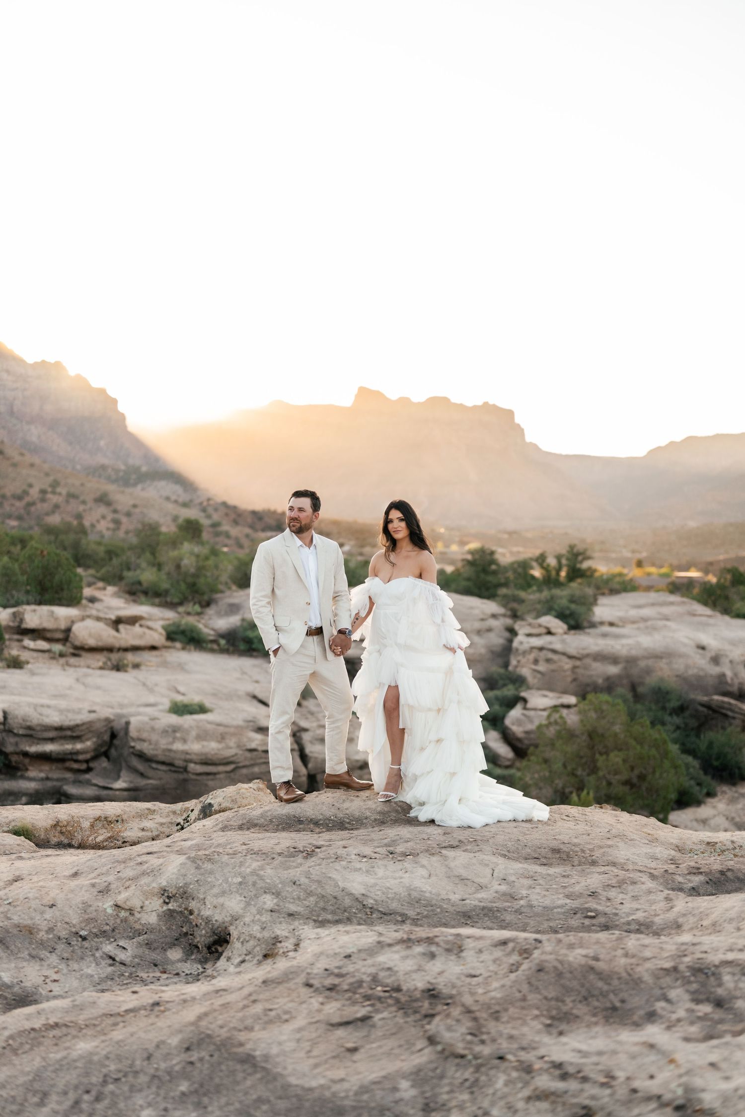 Zion destination photos. A couple in white wedding attire poses on scenic desert rocks during golden hour with mountains in the background.