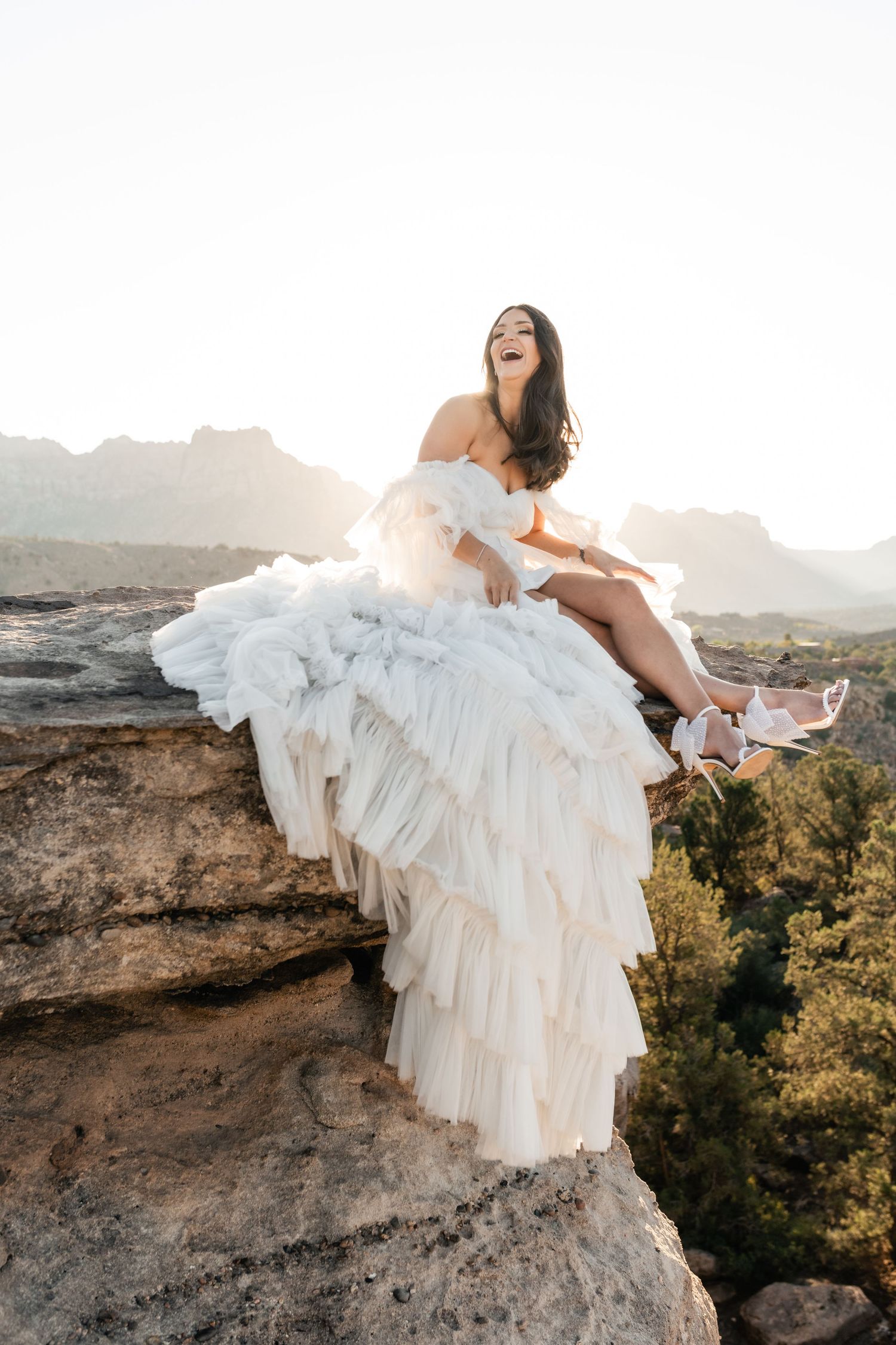 Bride in flowing white dress poses dramatically on desert cliff edge during sunset.