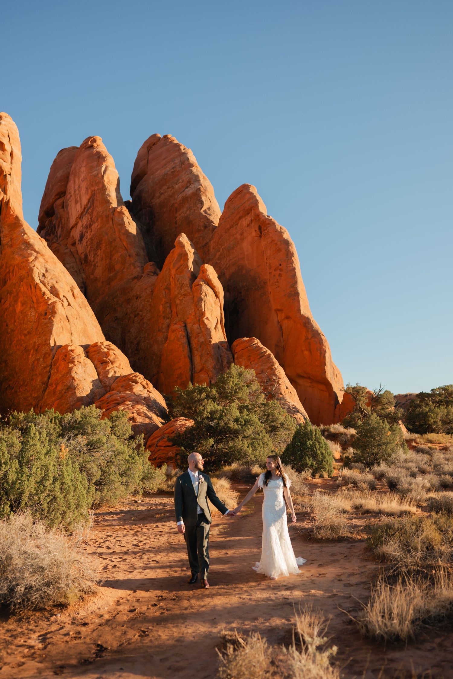 Two people walk together through a scenic desert landscape with towering red rock formations at Arches National Park.