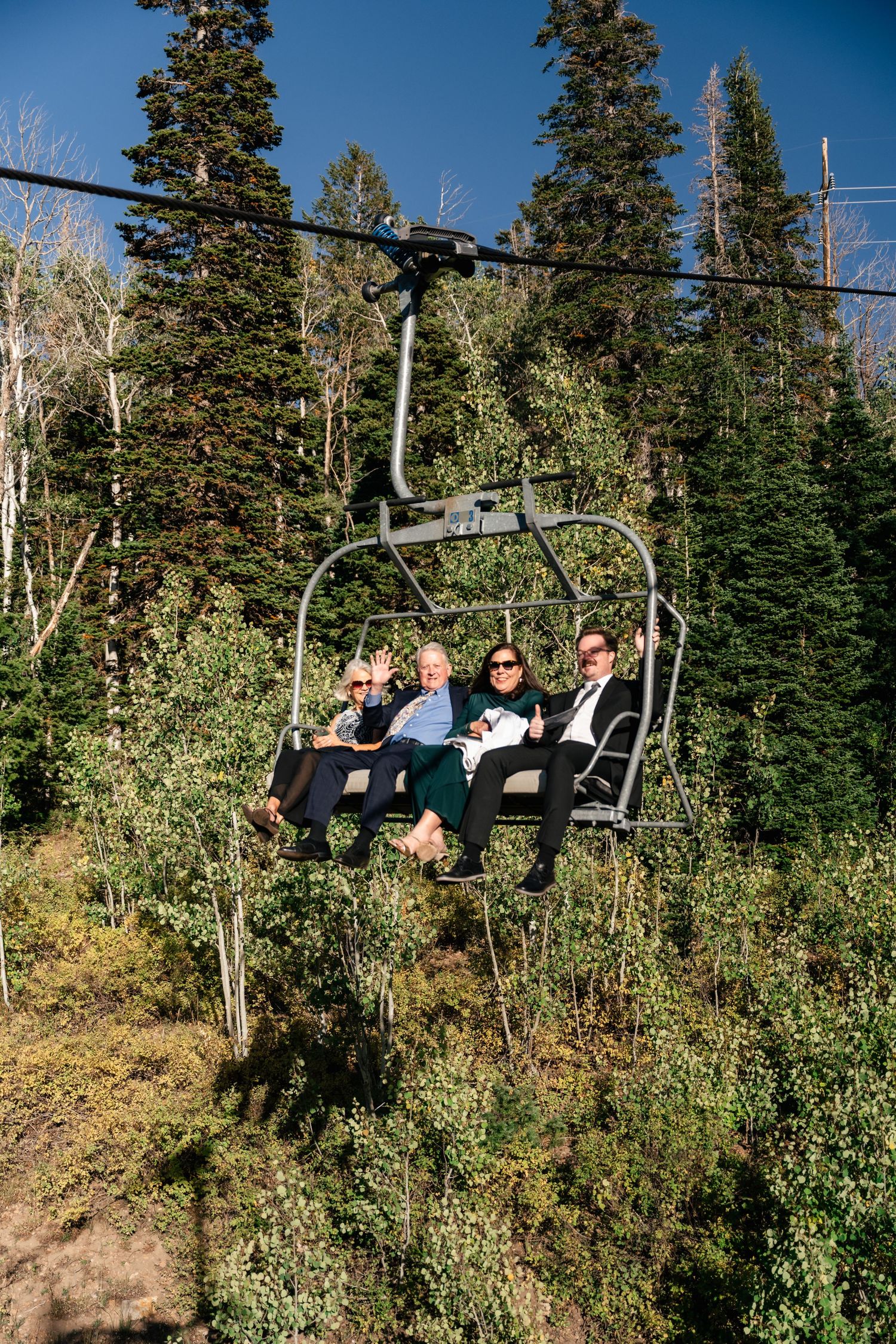 Wedding party enjoys a fun ride on a ski lift chair surrounded by evergreen trees in the mountains. Park City Wedding Photographer.