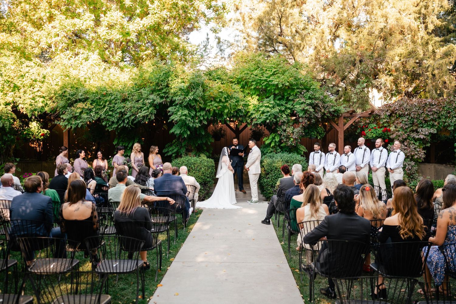 Outdoor wedding ceremony taking place in a garden setting with rows of chairs and a white aisle runner.