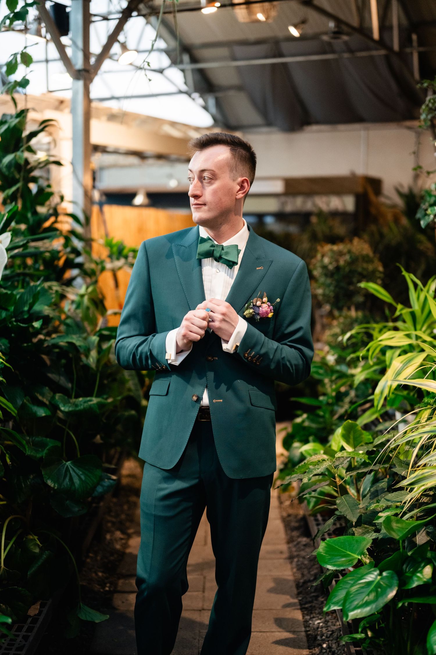 A groom in an emerald green suit and bow tie stands on a lush garden path surrounded by tropical plants.