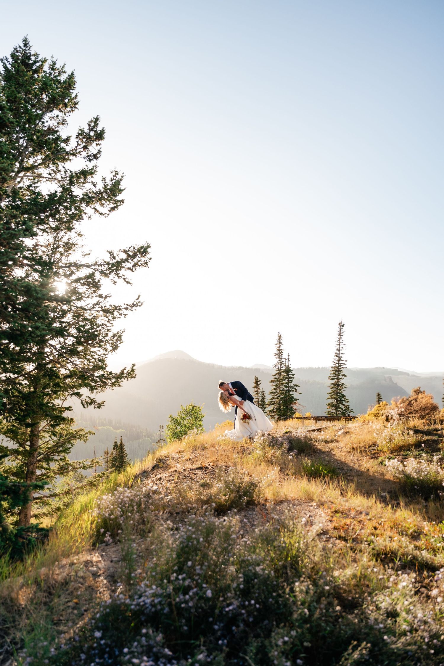 A couple shares a romantic moment on a mountain summit at sunset with evergreen trees and rolling hills in the backdrop. Utah Wedding Photo.