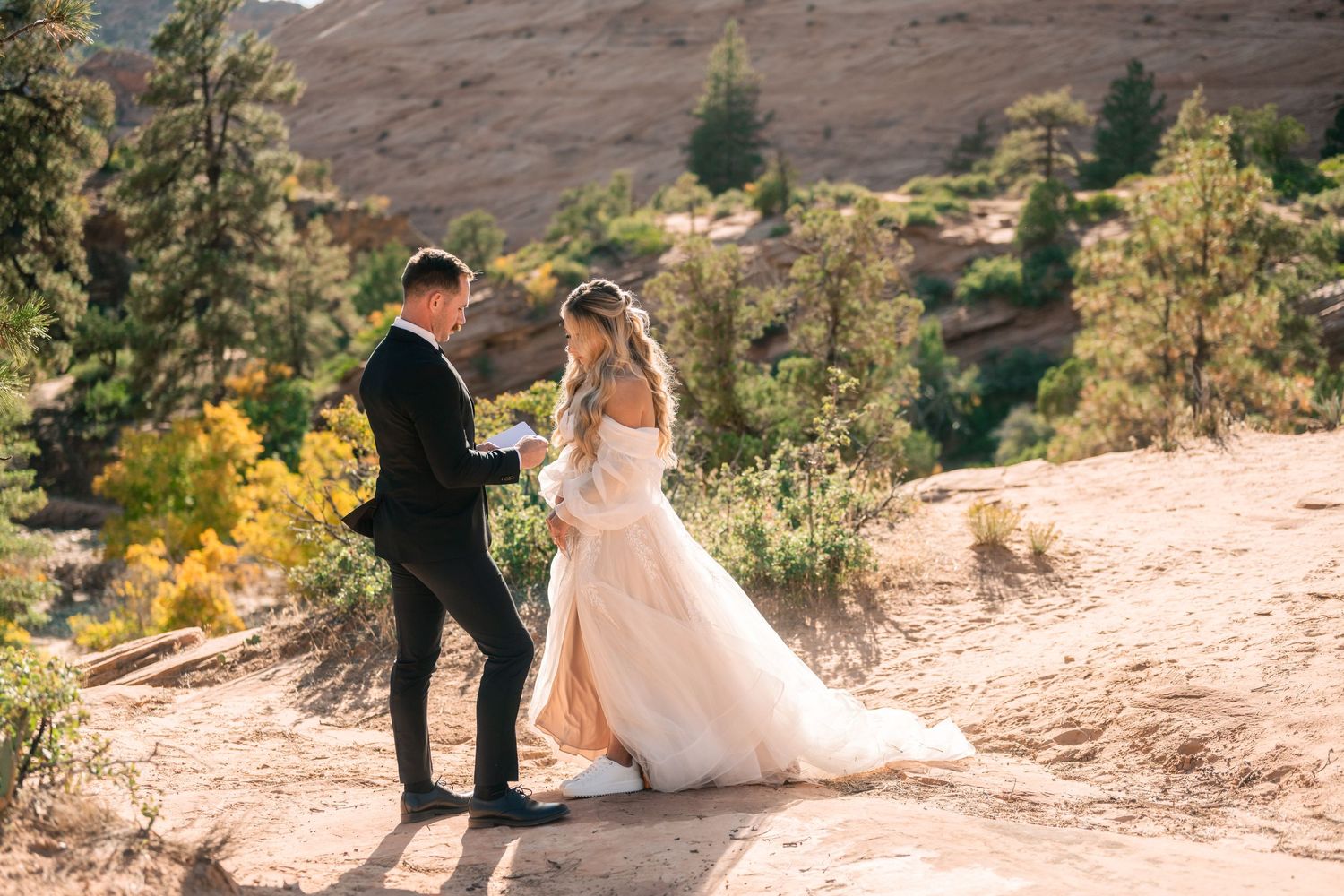 A wedding couple stands on a desert trail surrounded by pine trees and red rock formations on a sunny day.