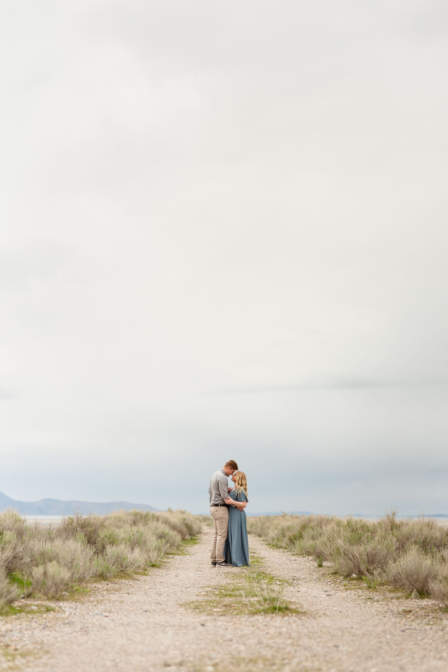 Two people embrace on a desert path under an overcast sky.