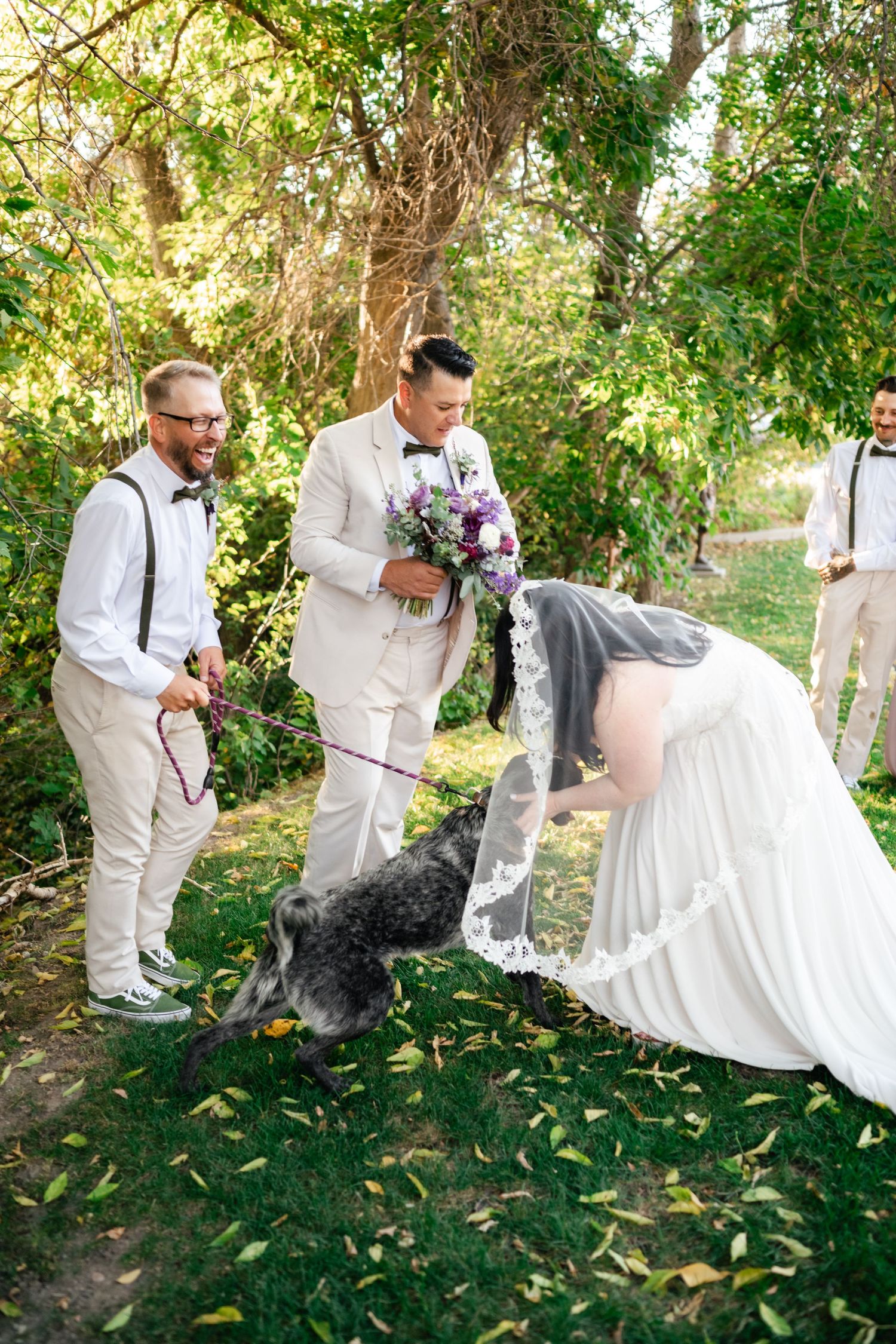 A wedding party in light colored attire stops to greet a large black dog on a tree-lined path during an outdoor ceremony.
