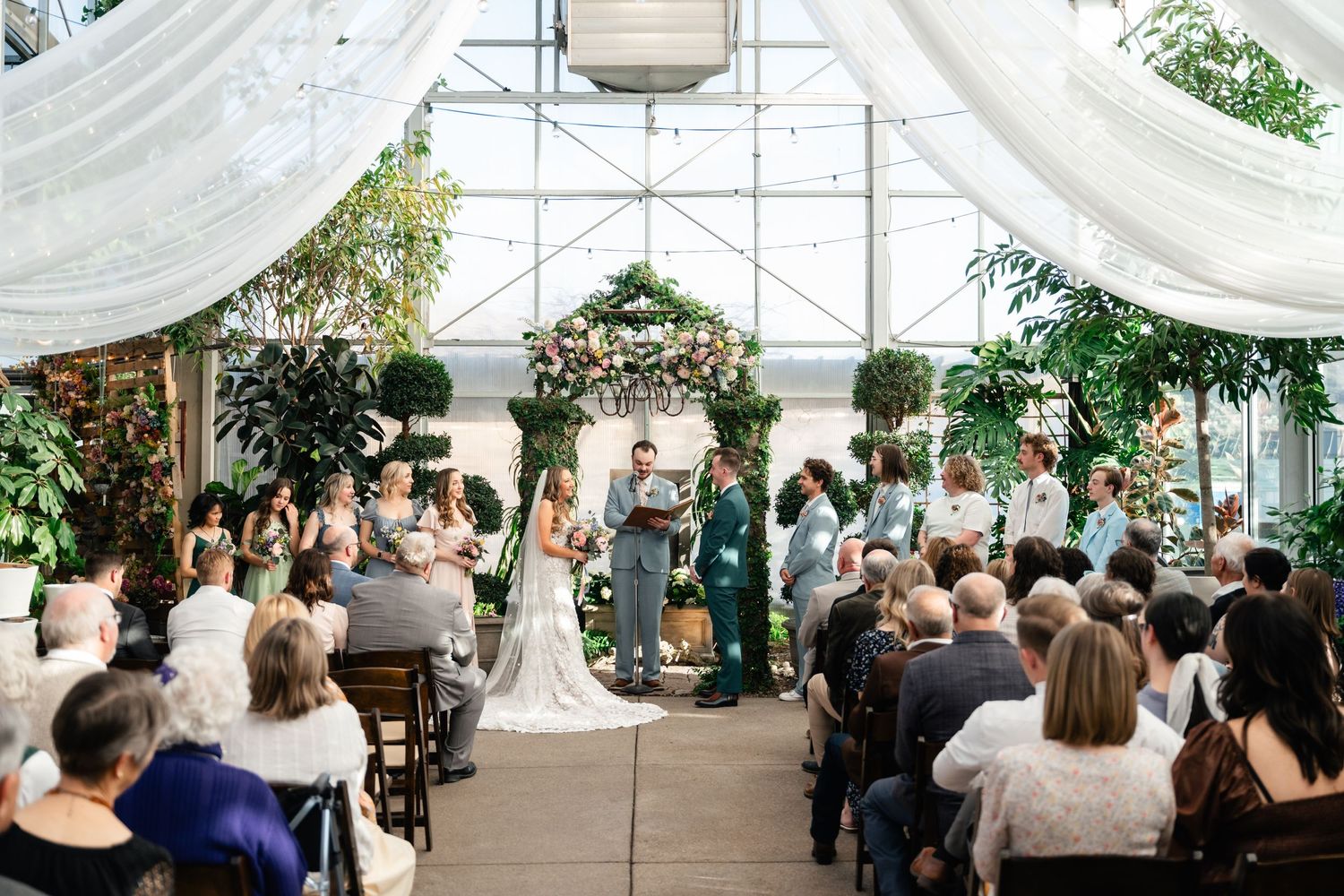 A wedding ceremony takes place in a bright conservatory filled with guests and greenery beneath flowing white drapes.