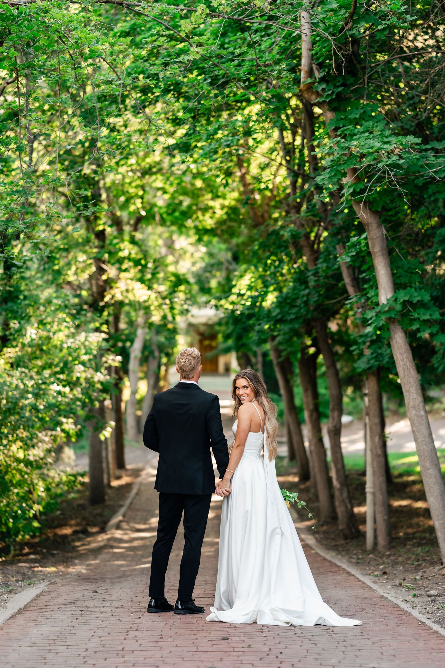 A couple walks along a tree-lined brick path in an elegant outdoor setting with dappled sunlight.