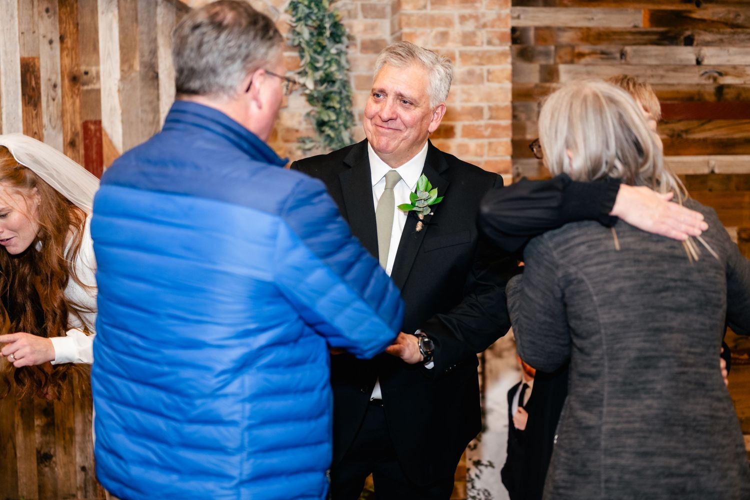 Guests in winter attire greet each other warmly at an indoor rustic venue with exposed brick walls.